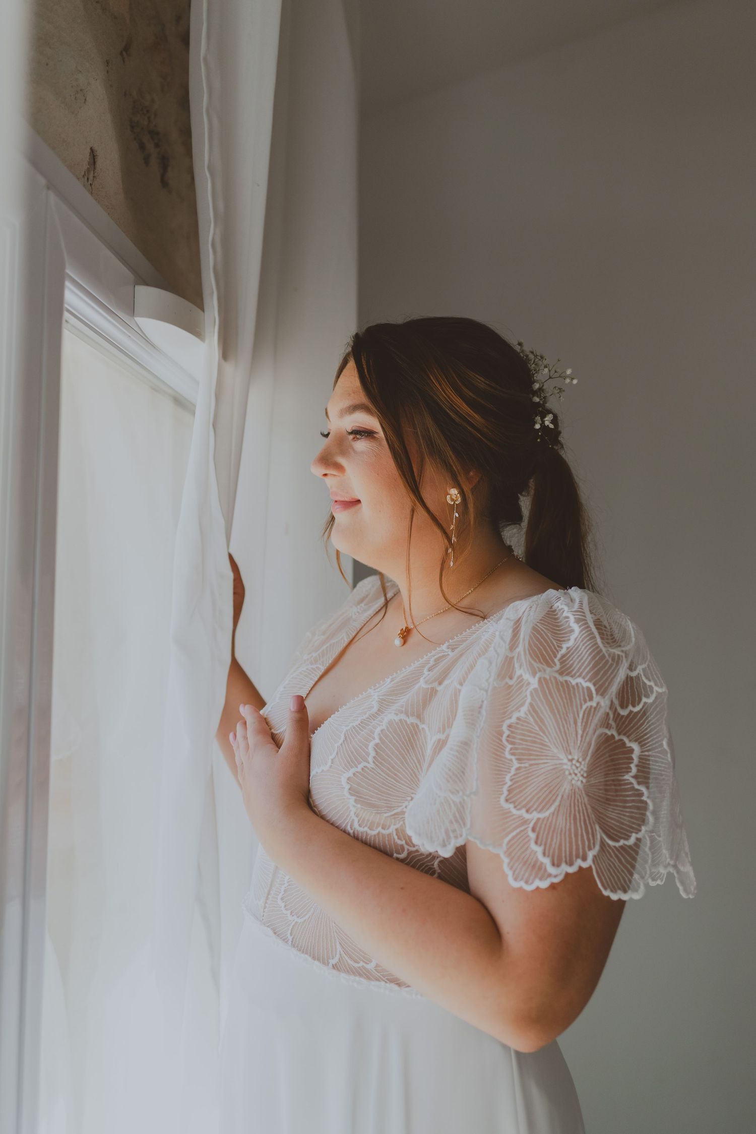Une mariée se tient à la fenêtre avec sa magnifique robe de mariée. Photo par Alice Enrico, photographe Mariage à Montpellier.