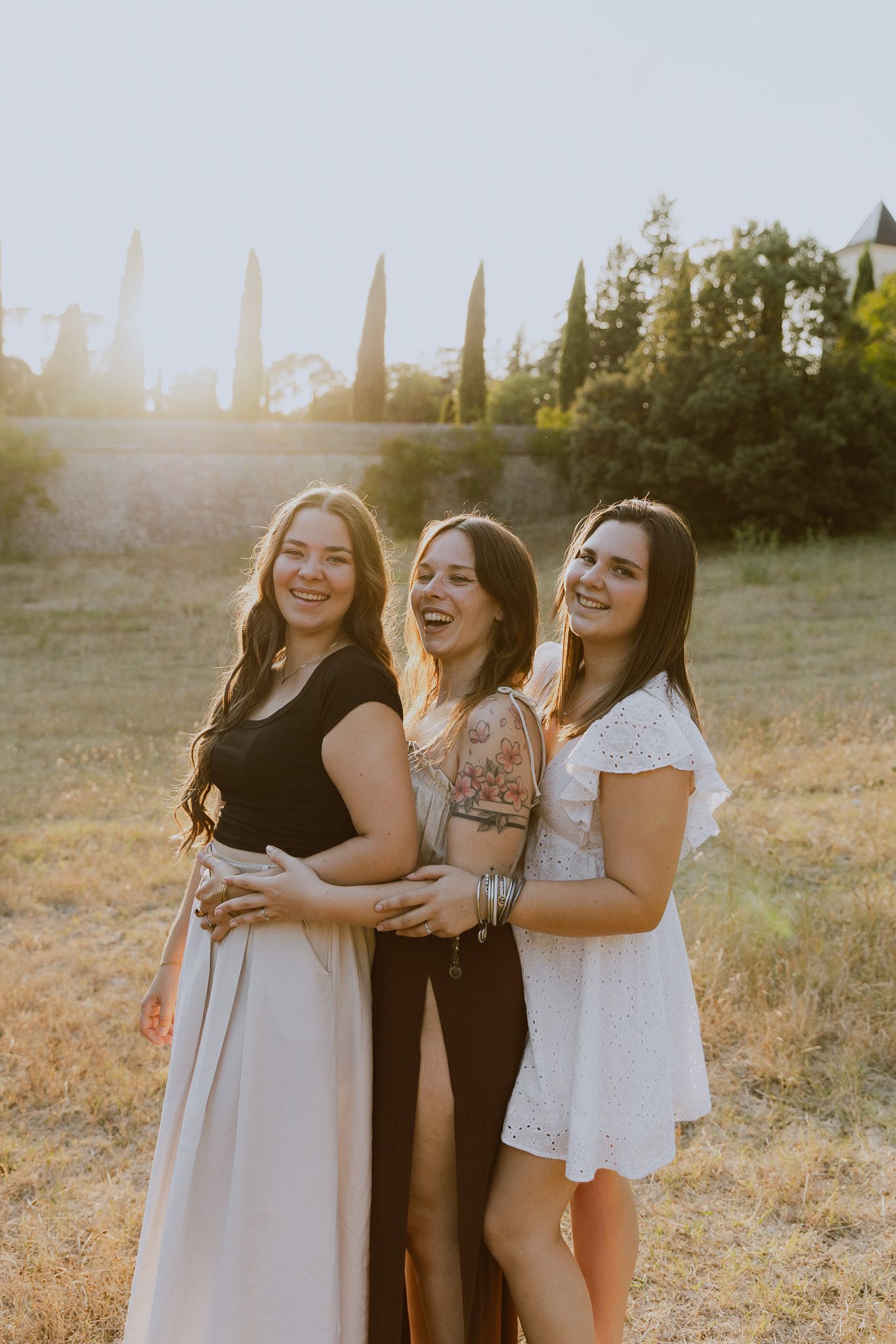 Three women laughing and embracing in a sunlit field with cypress trees in the background.