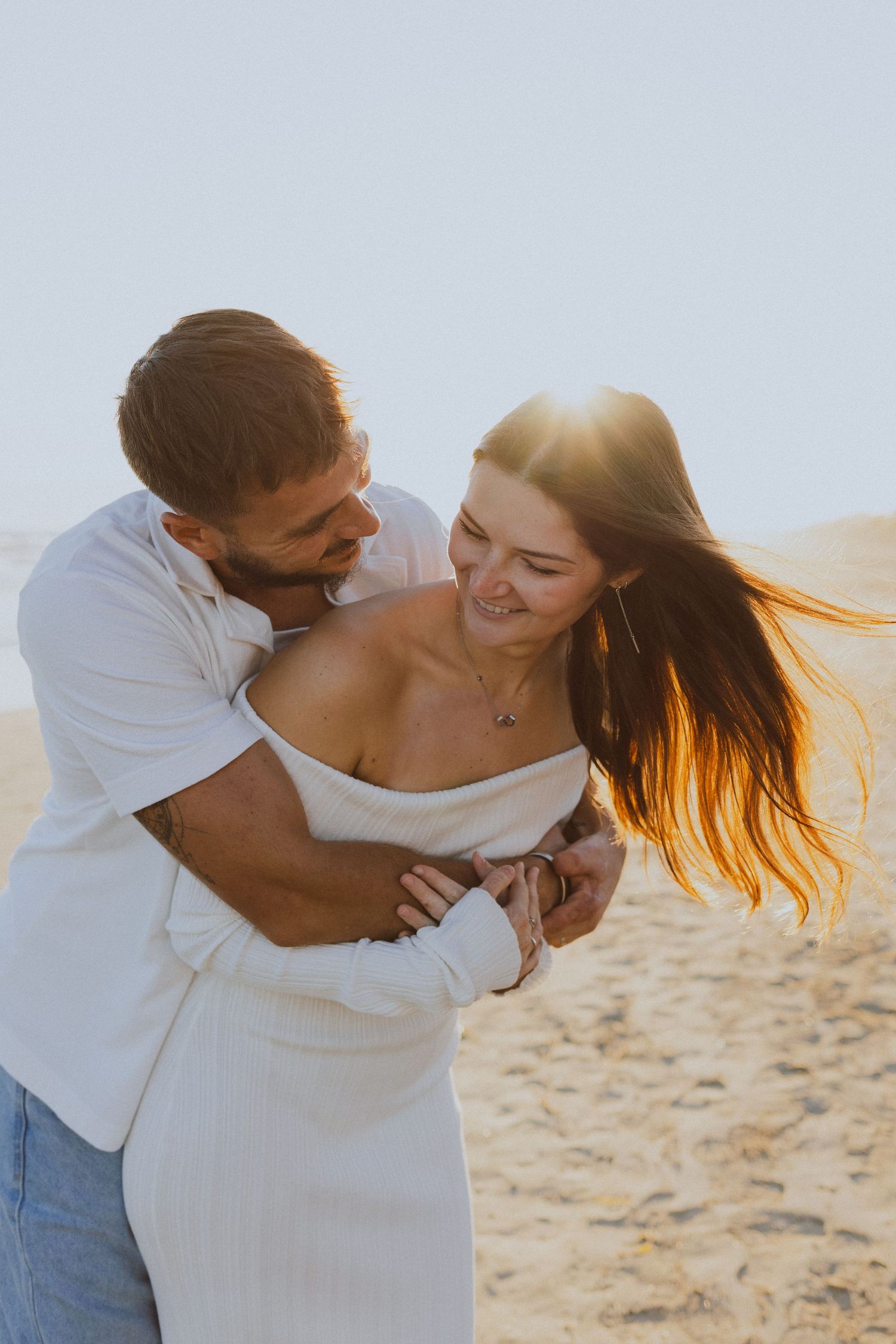 Un couple rigole ensemble sur le sable pendant une séance photo engagement, pendant la golden hour.