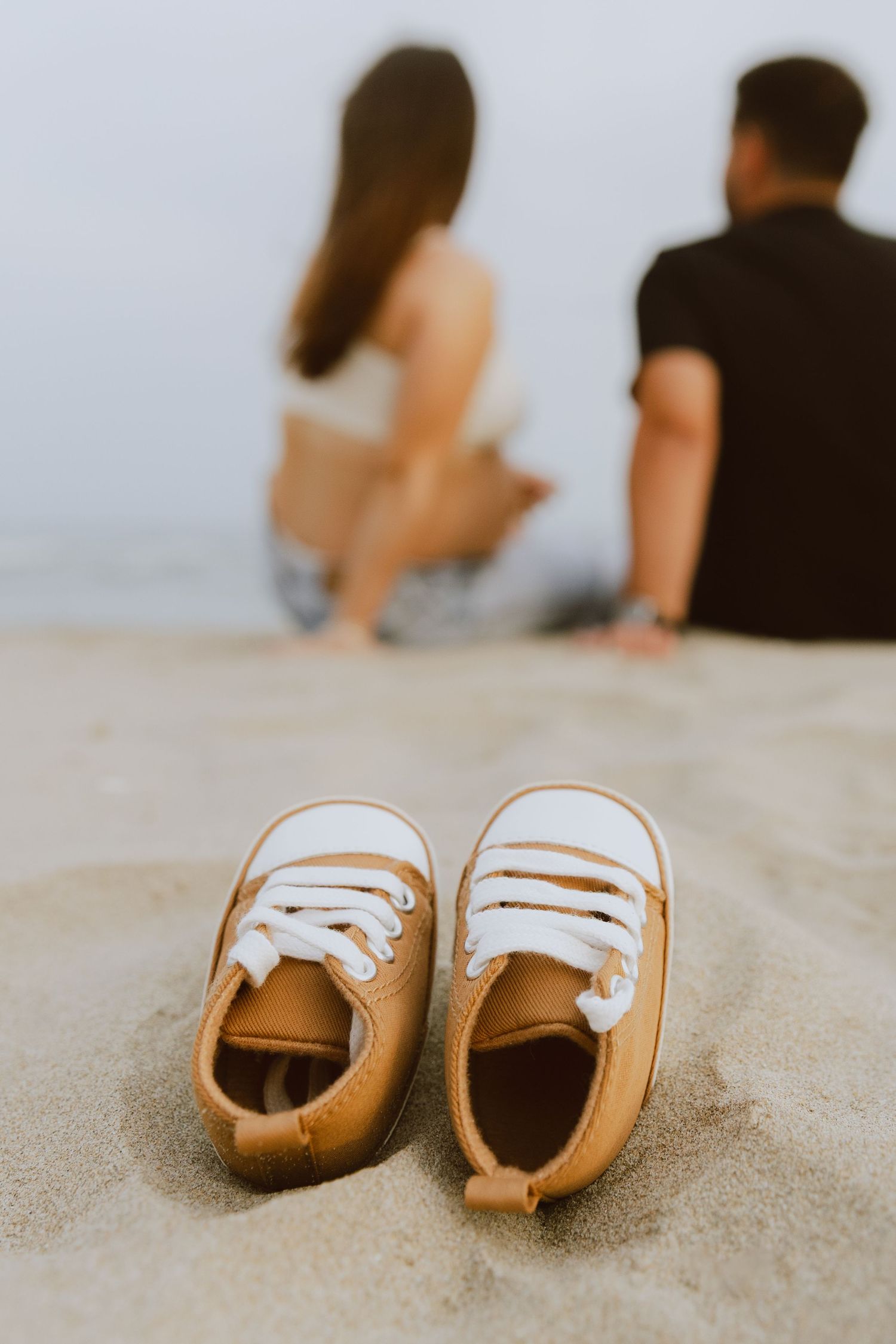 Des petites chaussures de bébé en premier plan, avec les futurs parents qui regardent la mer en second plan.