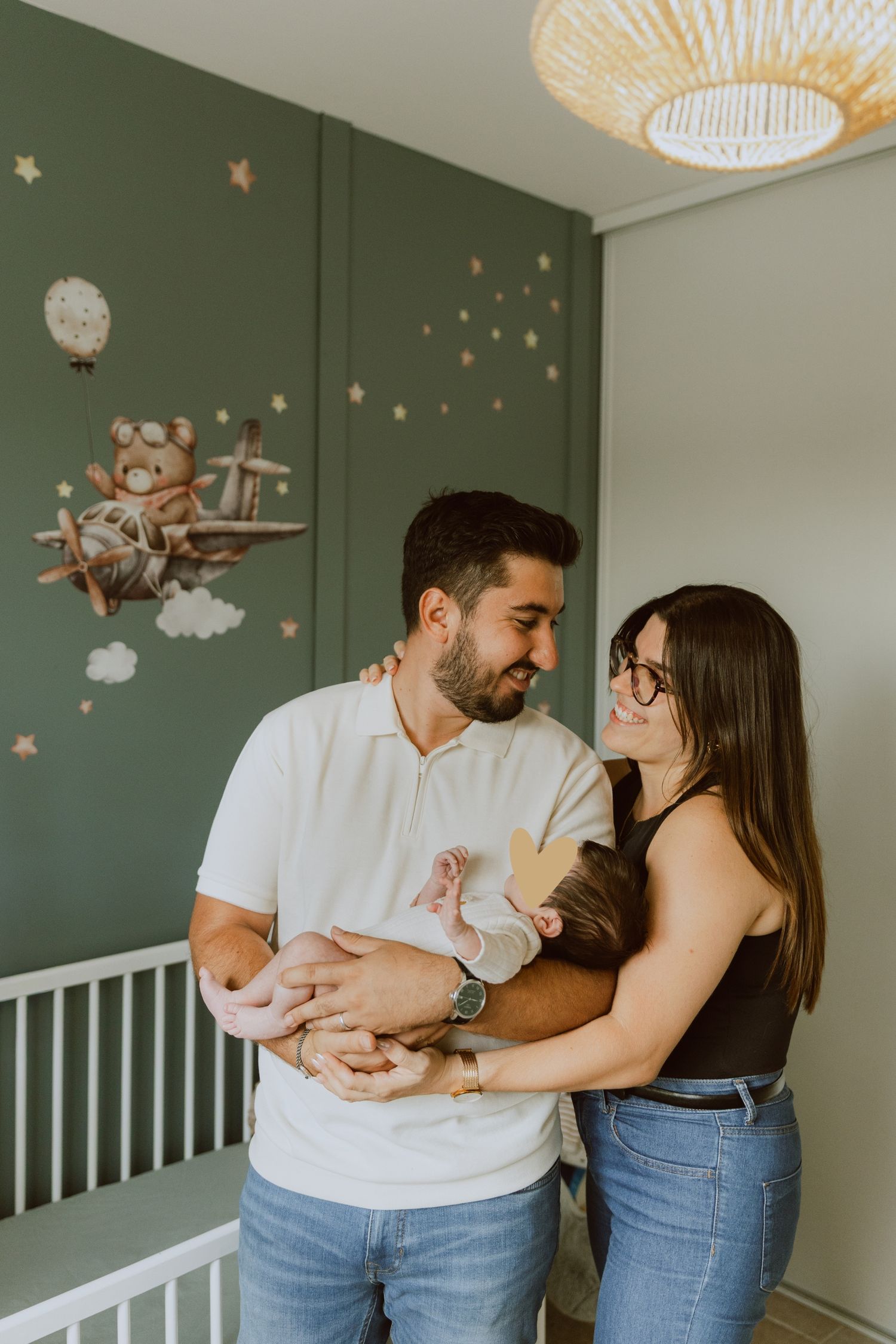 Shooting photo famille à domicile, les jeunes parents portent leur bébé dans sa chambre d'enfant vert sauge
