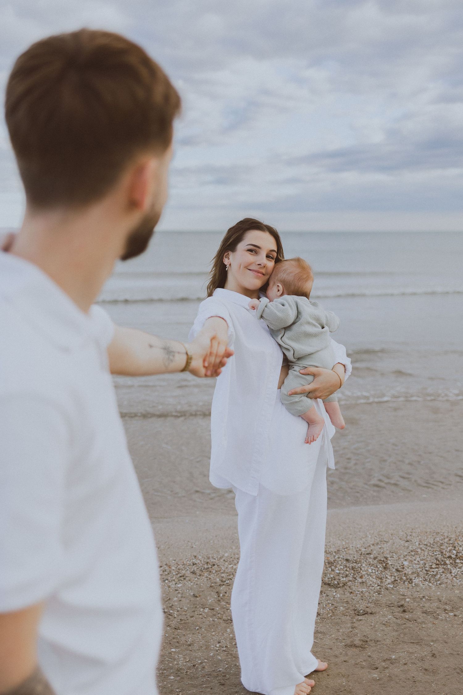Une jeune maman regarde le papa, en tenant son bébé, au bord de la mer.
