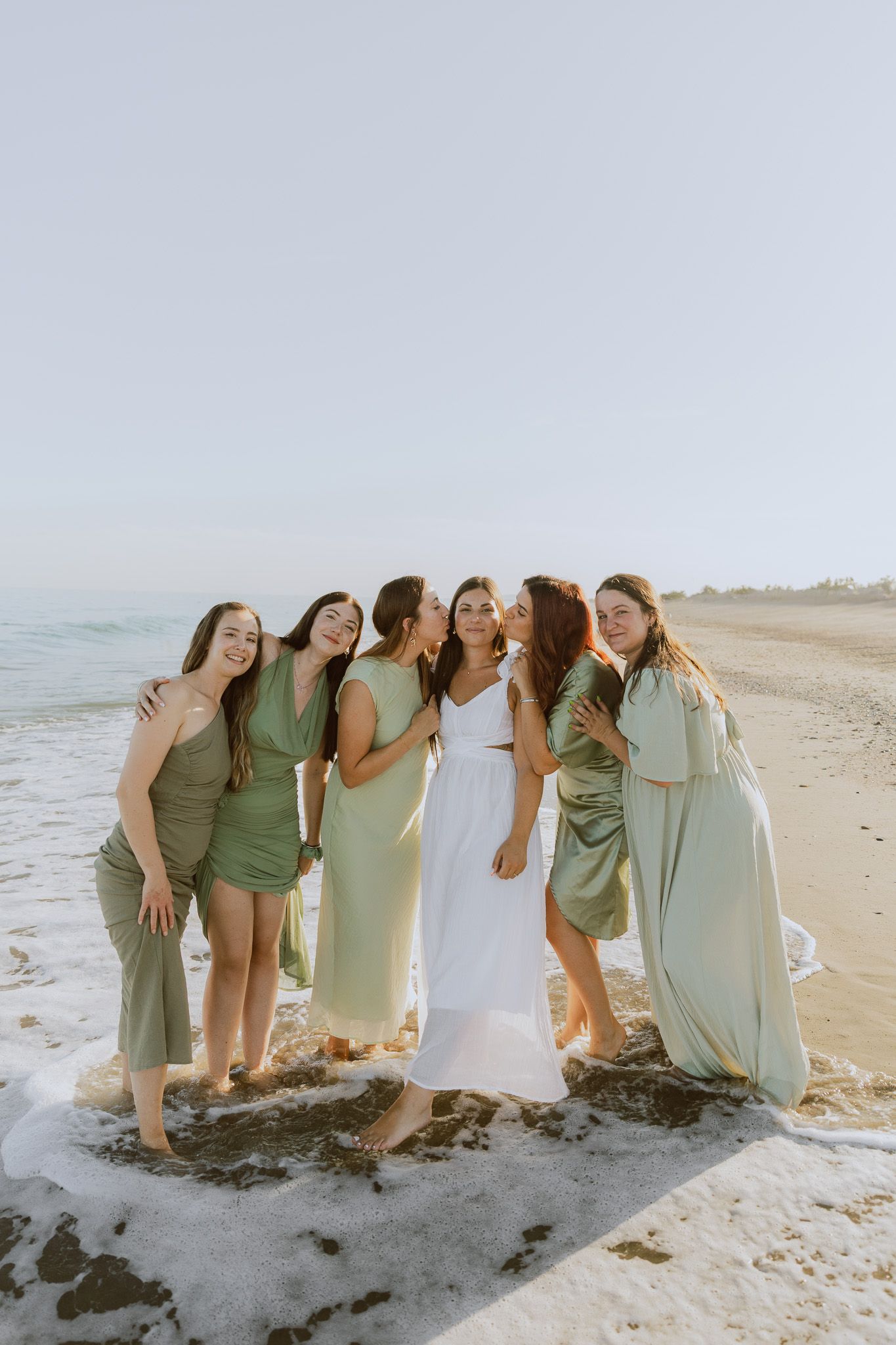 Un groupe d'amies fête le EVJF d'une copine, elles rigolent au bord de la mer, toutes habillées avec des robes en satin vert sauge.