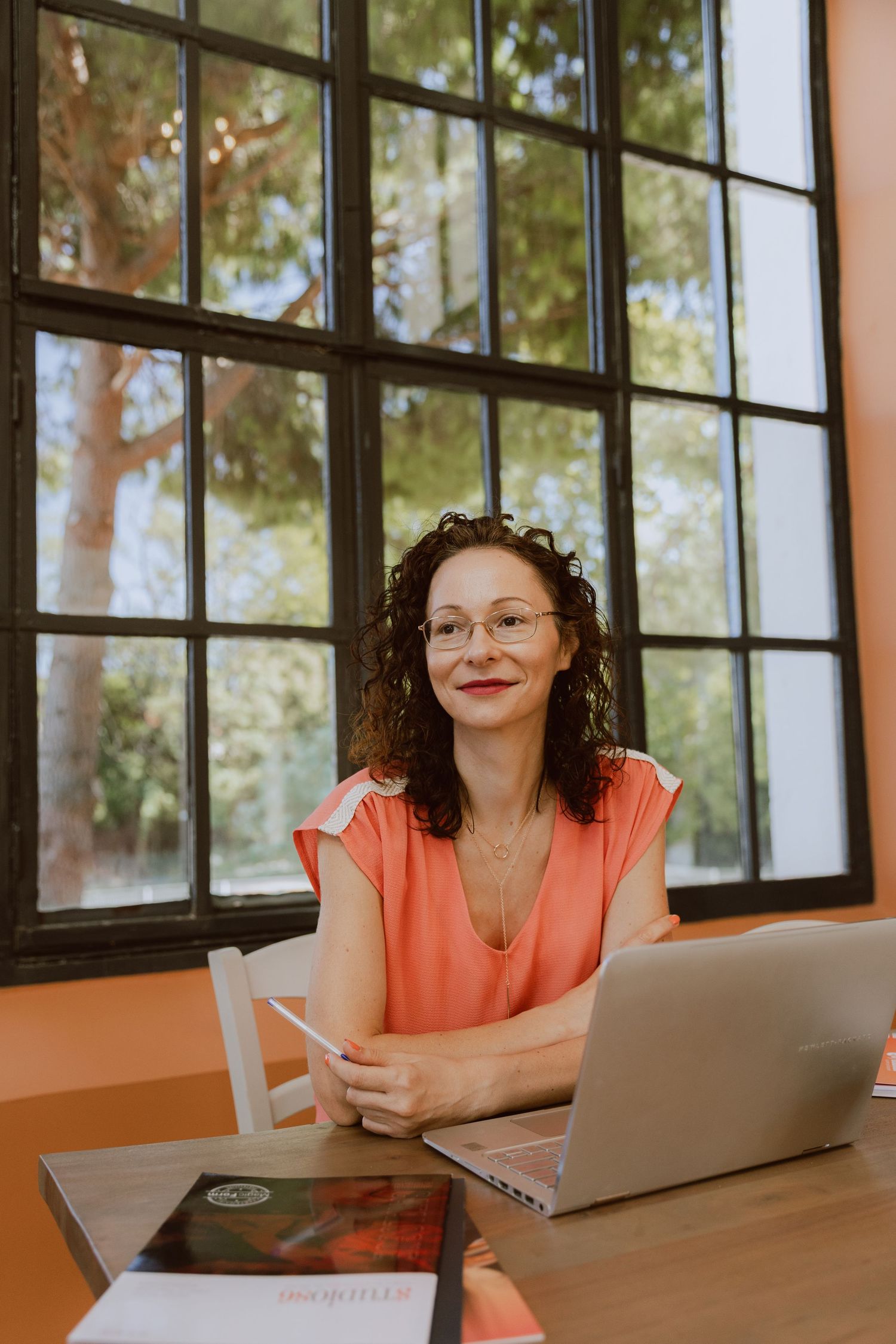 Femme entrepreneuse assise sur son bureau avec son ordinateur, devant une belle et grande fenêtre qui donne sur un arbre.