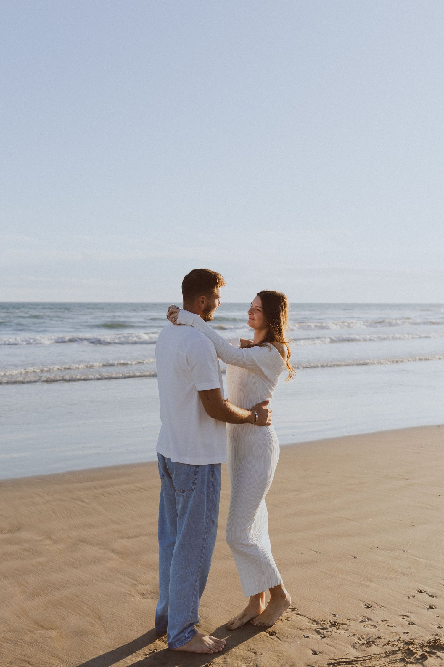 Un couple se regarde au bord de l'eau à la plage de Montpellier pendant leur shooting photo engagement à la golden hour.