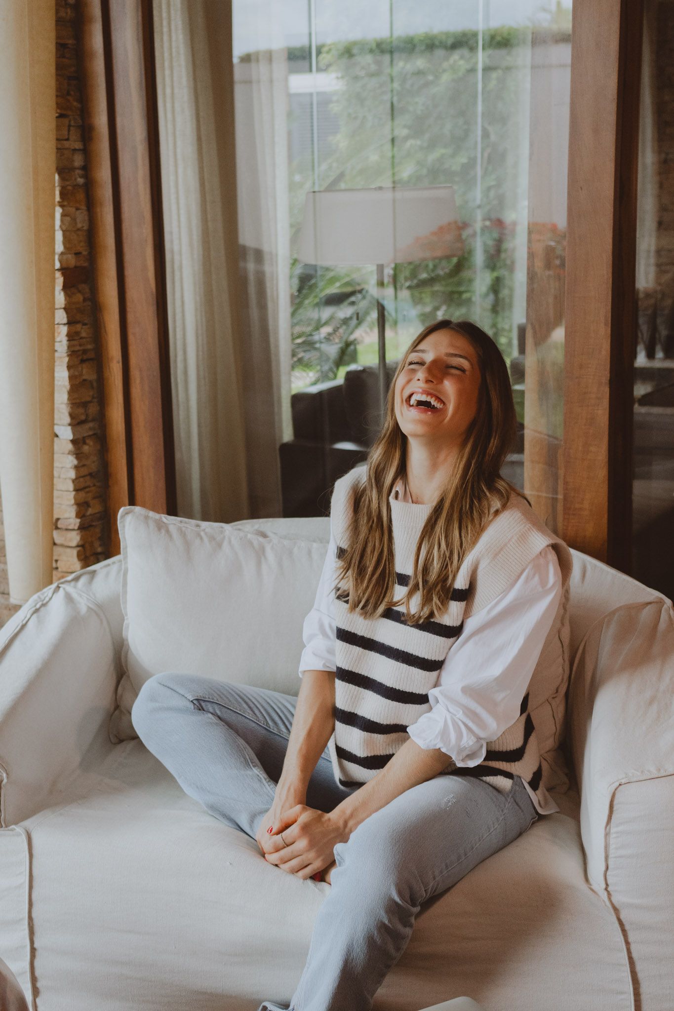 Jeune femme assise sur un canapé blanc, qui rigole de façon heureuse dans un salon lumineux et bien décoré.