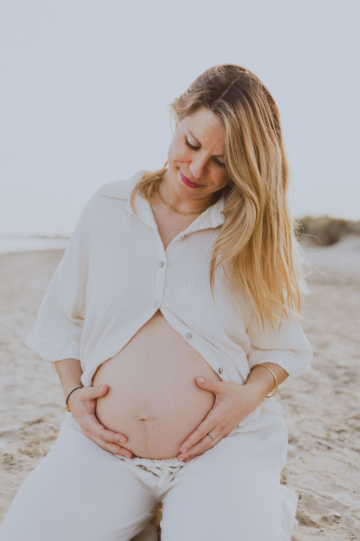 Une femme enceinte, habillée en blanc, regarde son ventre rond avec tendresse. Elle est assise sur la plage.