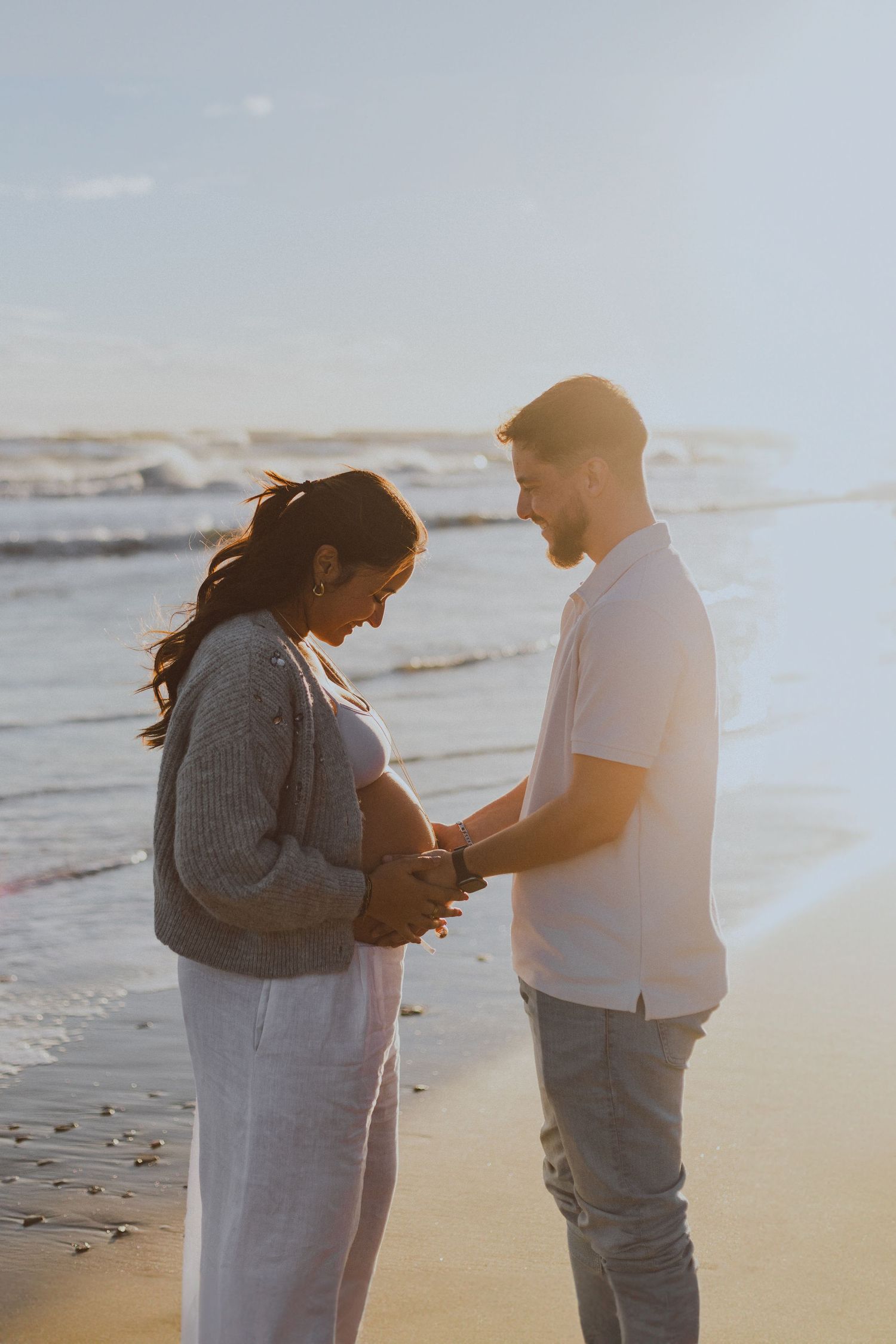 Un couple se regarde au bord de l'eau à la plage de Montpellier pendant leur shooting photo de grossesse à la golden hour.