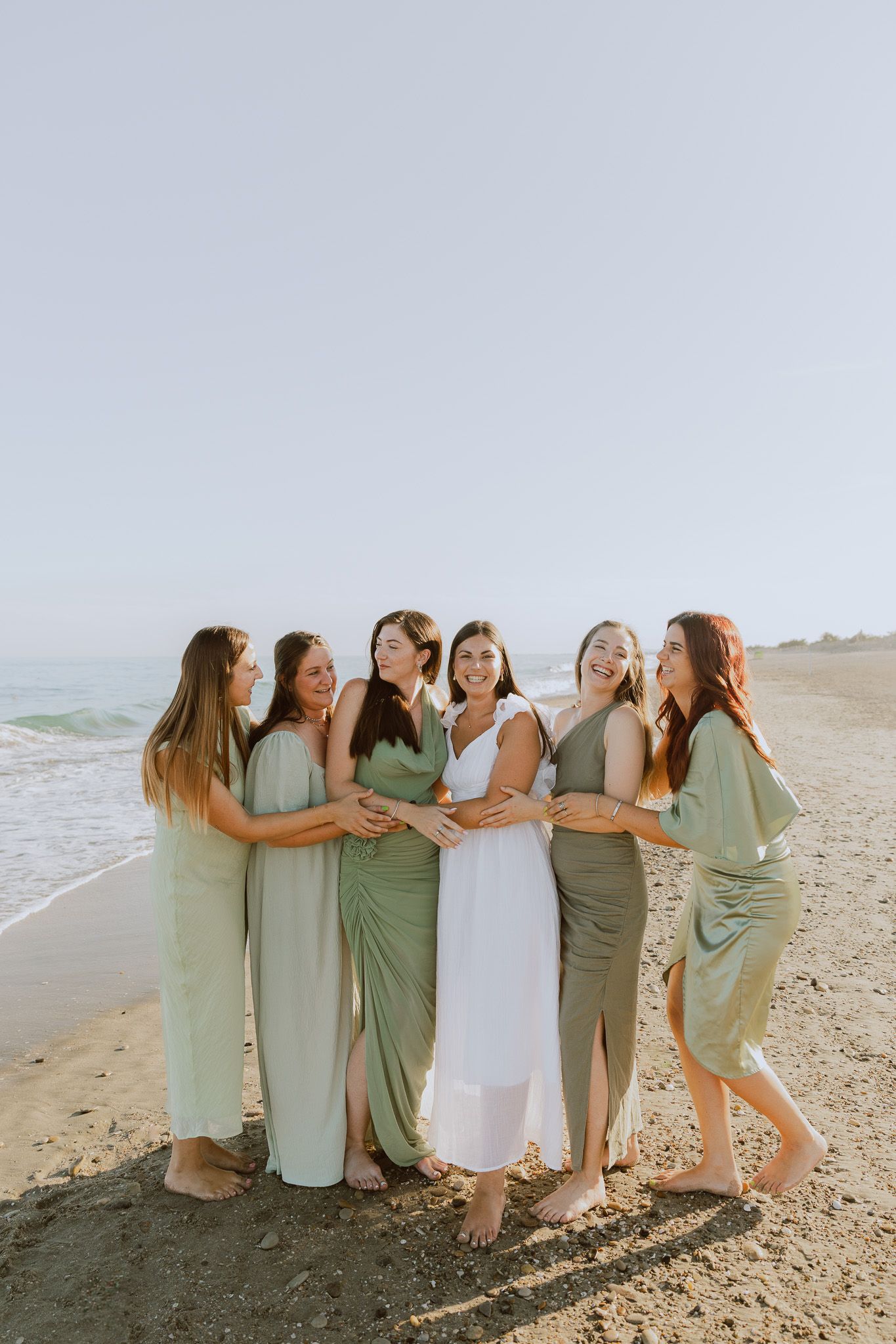 Un groupe d'amies fête le EVJF d'une copine, elles rigolent au bord de la mer, toutes habillées avec des robes en satin vert sauge.