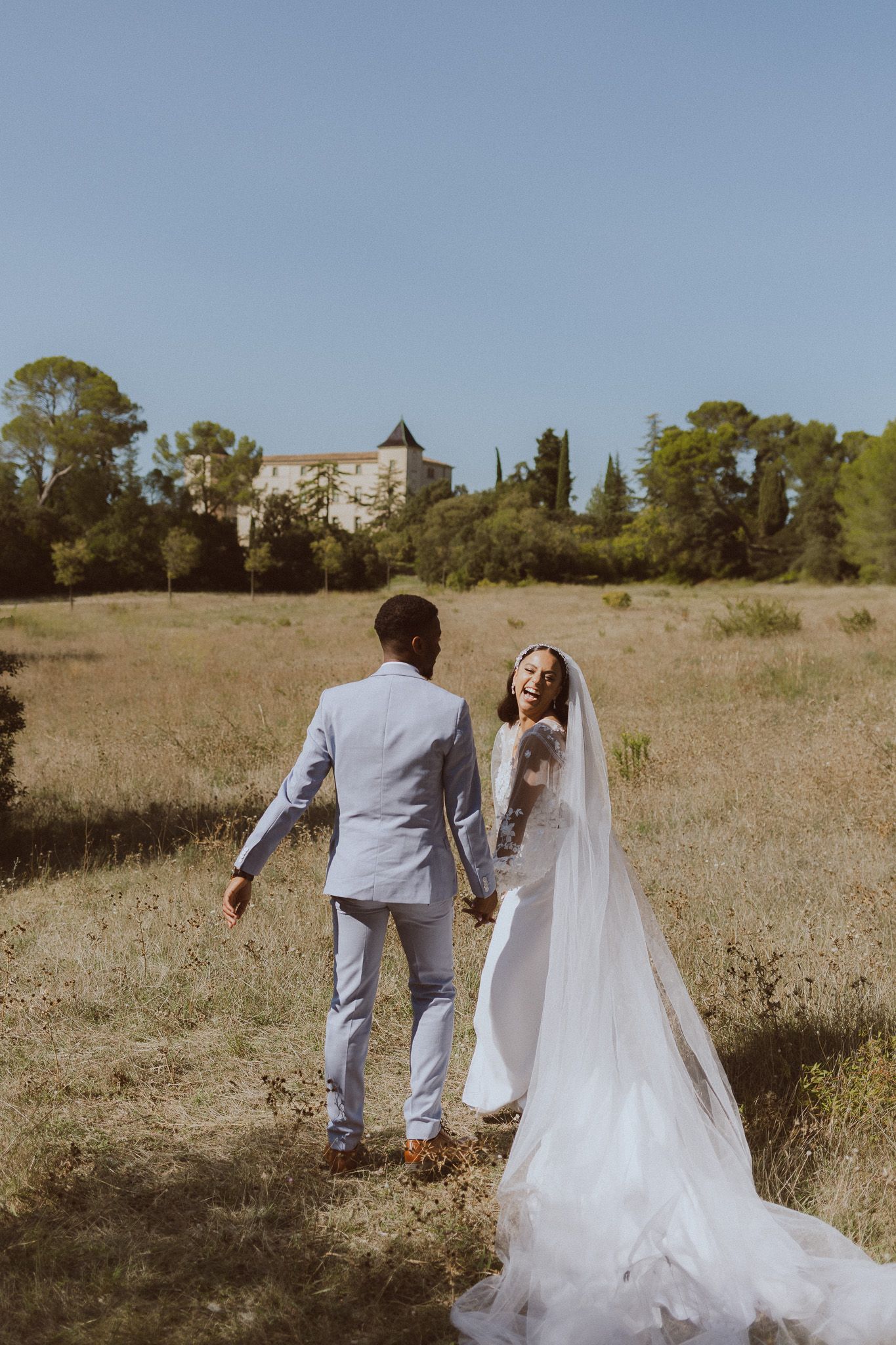 Un couple de mariés rigole dans un champ devant un château. Photo par Alice Enrico, photographe mariage à Montpellier.