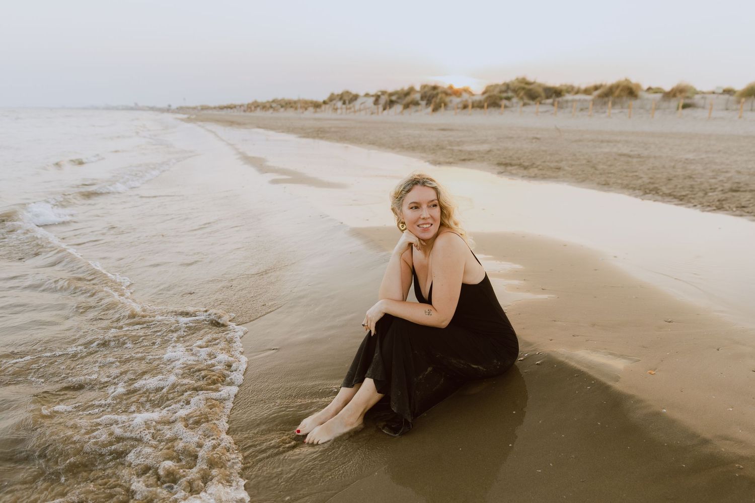 Une femme en robe noire assise au bord de l'eau, sur une plage, au coucher de soleil.