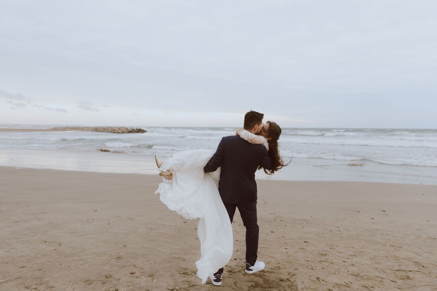 Un couple se mariés s'embrasse sur la plage dans un temps gris. Photo par Alice Enrico, photographe mariage à Montpellier.