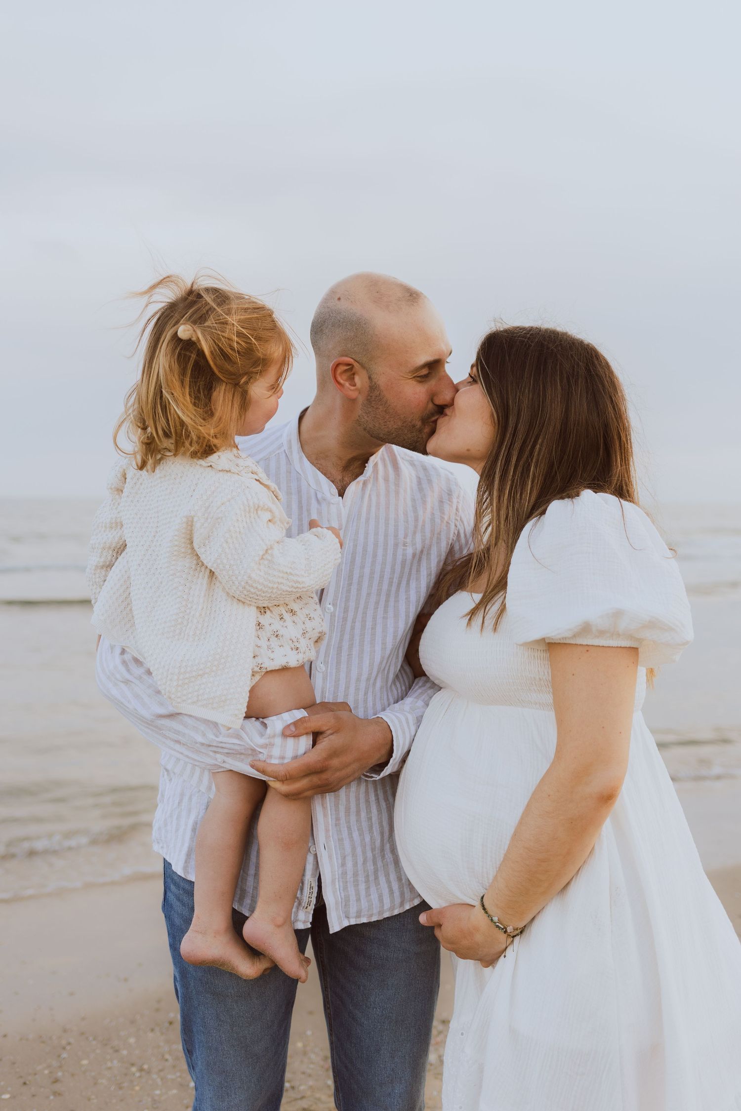 Des parents avec un enfant et qui attendent un bébé s'embrassent sur la plage pendant une séance photo de famille avec une photographe pro.