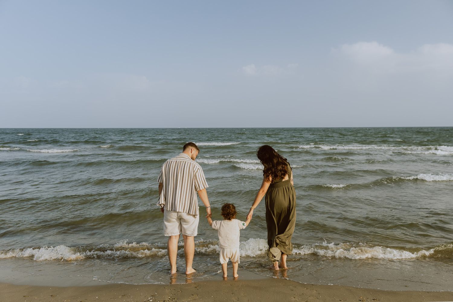 Des parents tiennent la main de leur petit enfant face à la mer, pendant une séance photo de famille avec une photographe pro.