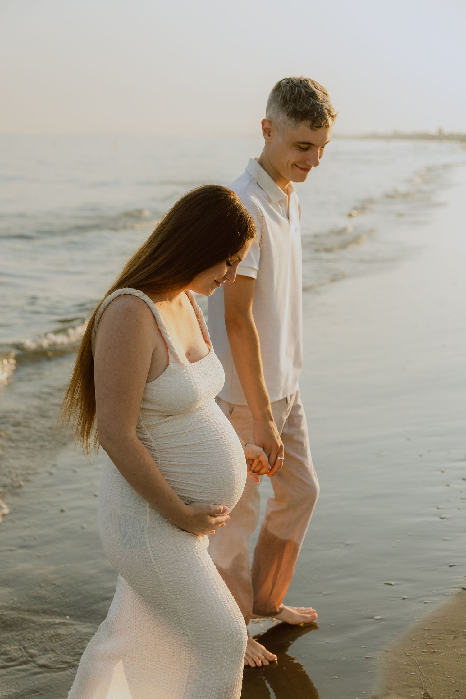 Un couple marche ensemble dans l'eau à la plage de Frontignan pendant leur séance photo de grossesse à la golden hour.