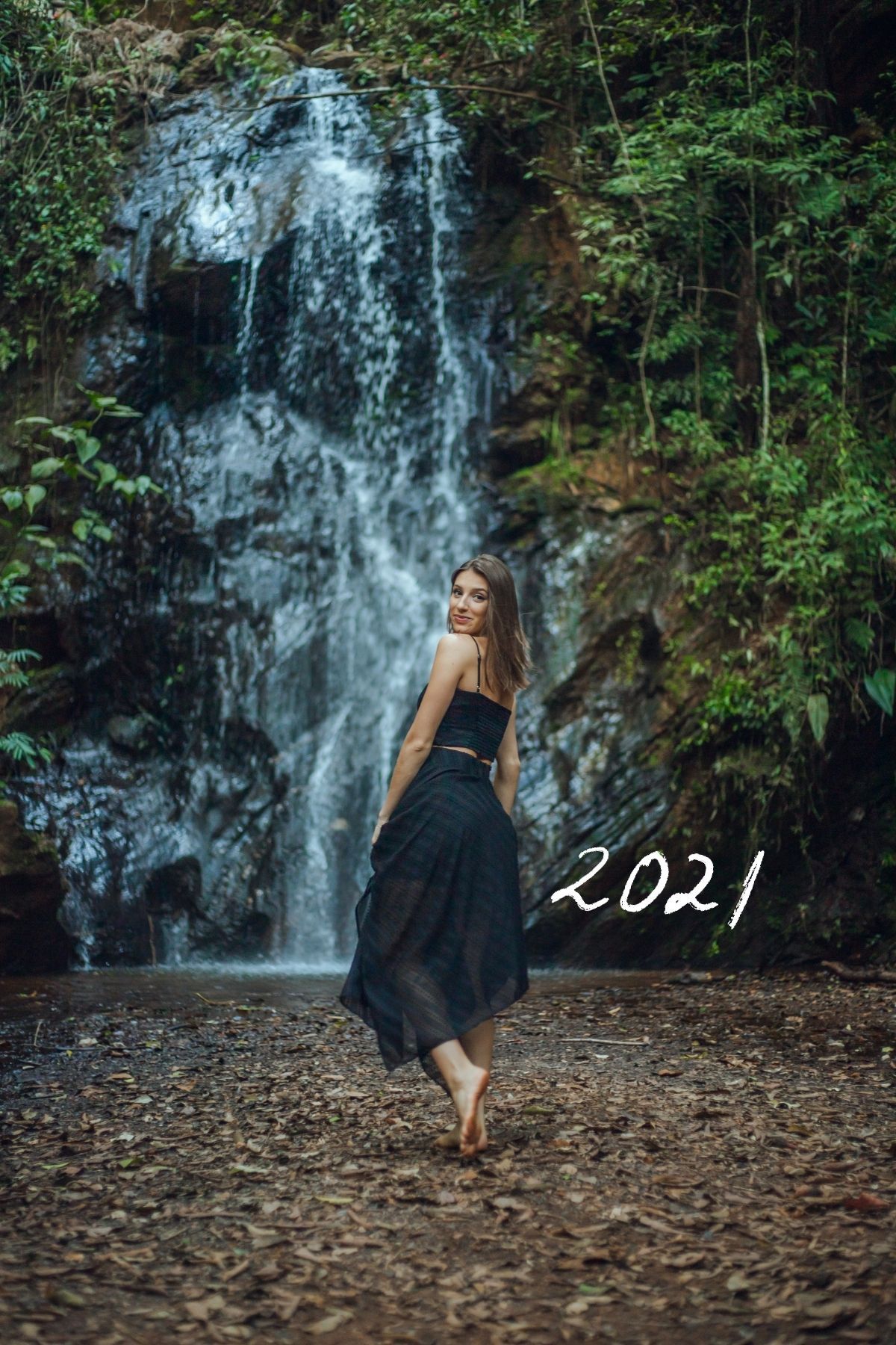 Jeune femme en robe noire marche vers une cascade dans un décor tropical.