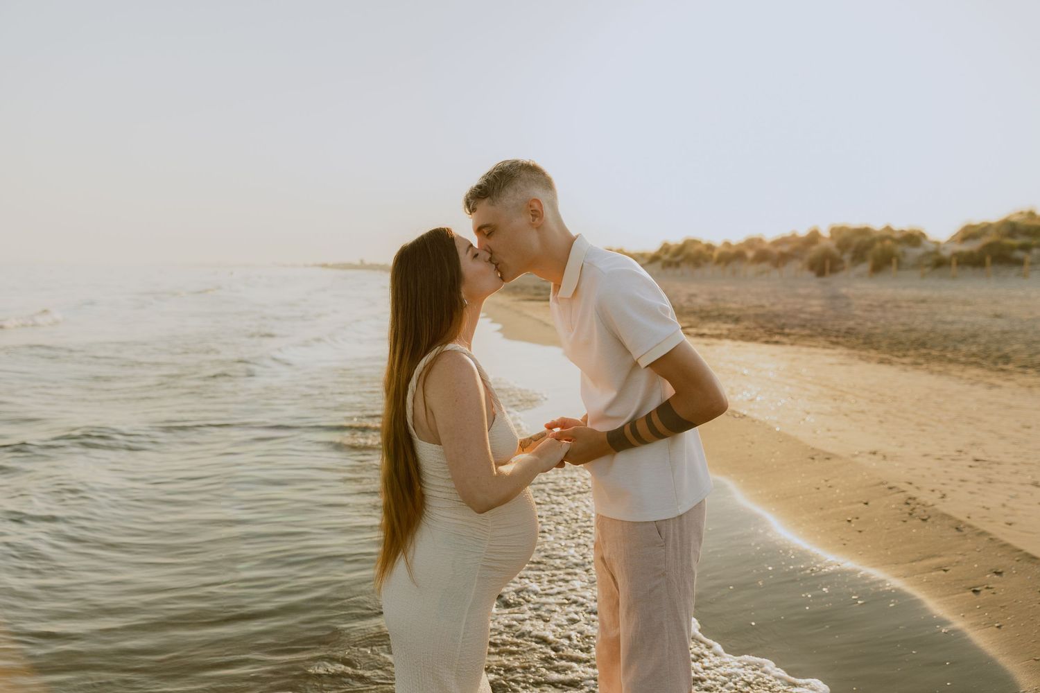 Un couple s'embrasse au bord de l'eau à la plage de Frontignan pendant leur séance photo de grossesse à la golden hour.