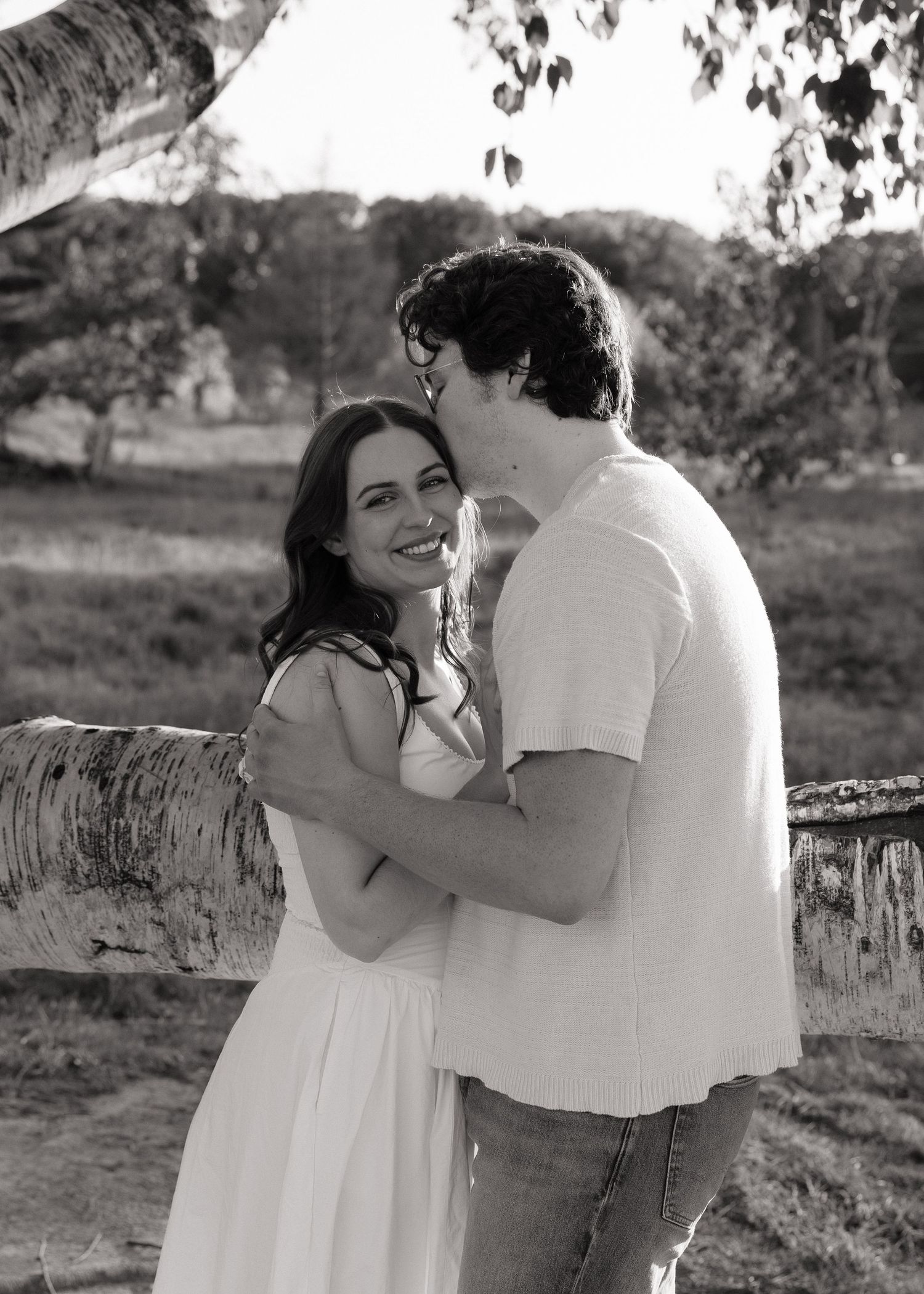 Black and white photo of a smiling woman in white dress being kissed on cheek by a man outdoors.