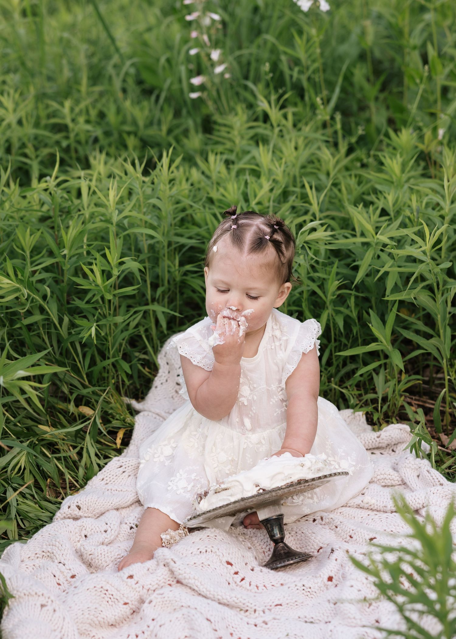 Toddler girl in white lace dress eating cake on blanket surrounded by lush green field plants.