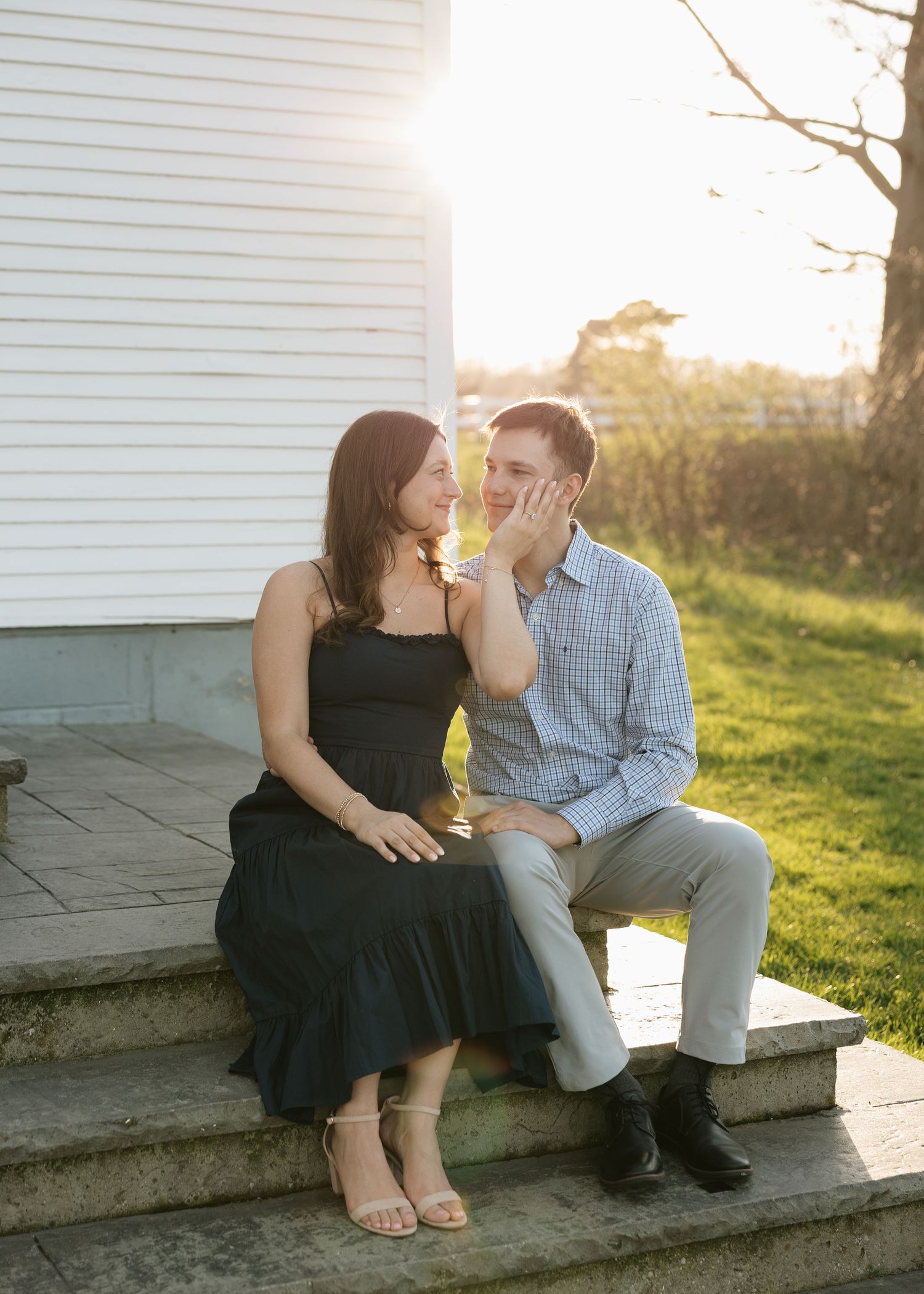 Couple sitting on stone steps at golden hour, woman in black dress touching man's face, sharing a romantic moment outdoors.