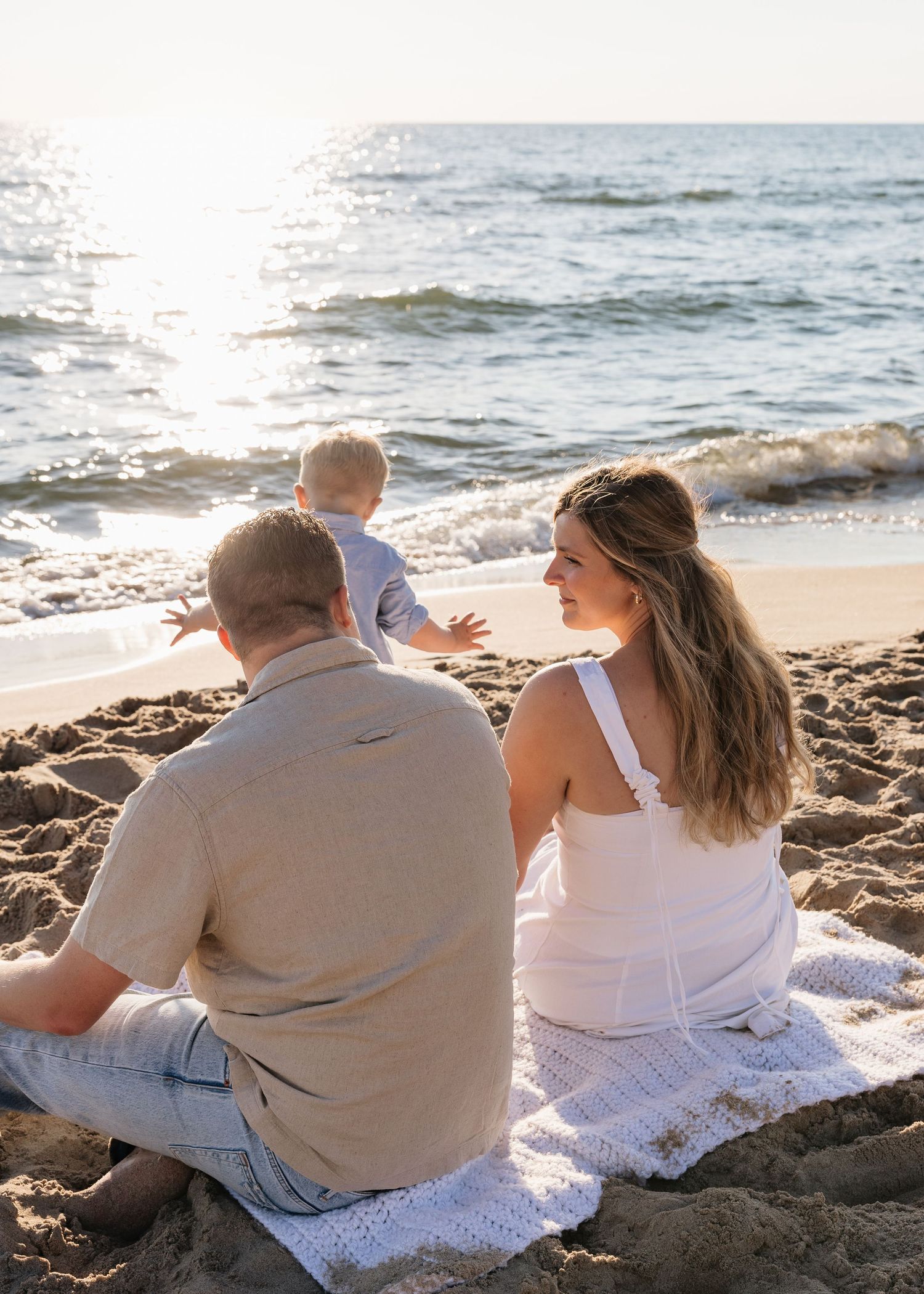 Family sitting on a blanket on a sandy beach at sunset, watching a toddler near the ocean waves.