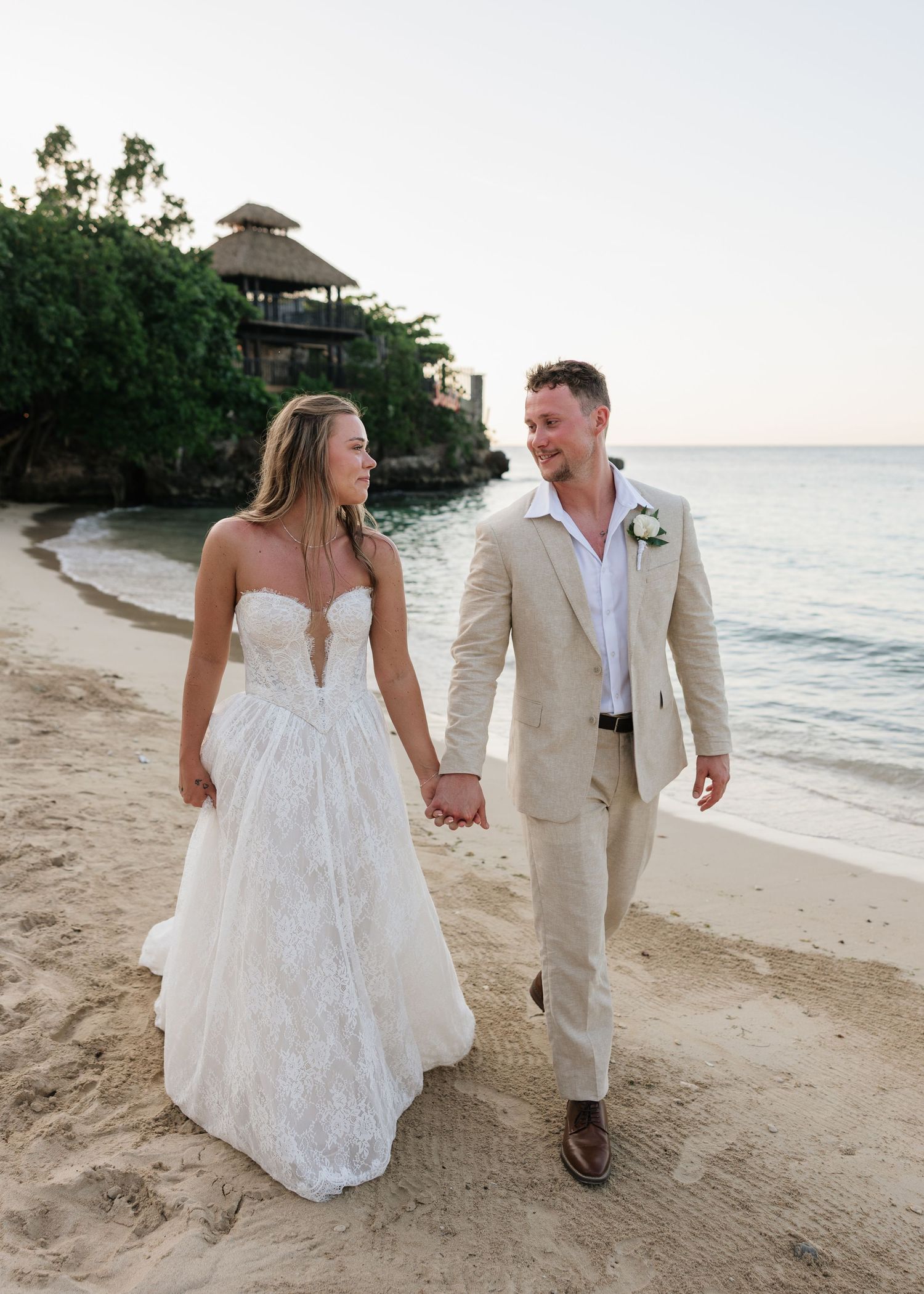Bride and groom walk hand-in-hand on a tropical beach at sunset, she in a white gown, he in a tan suit.