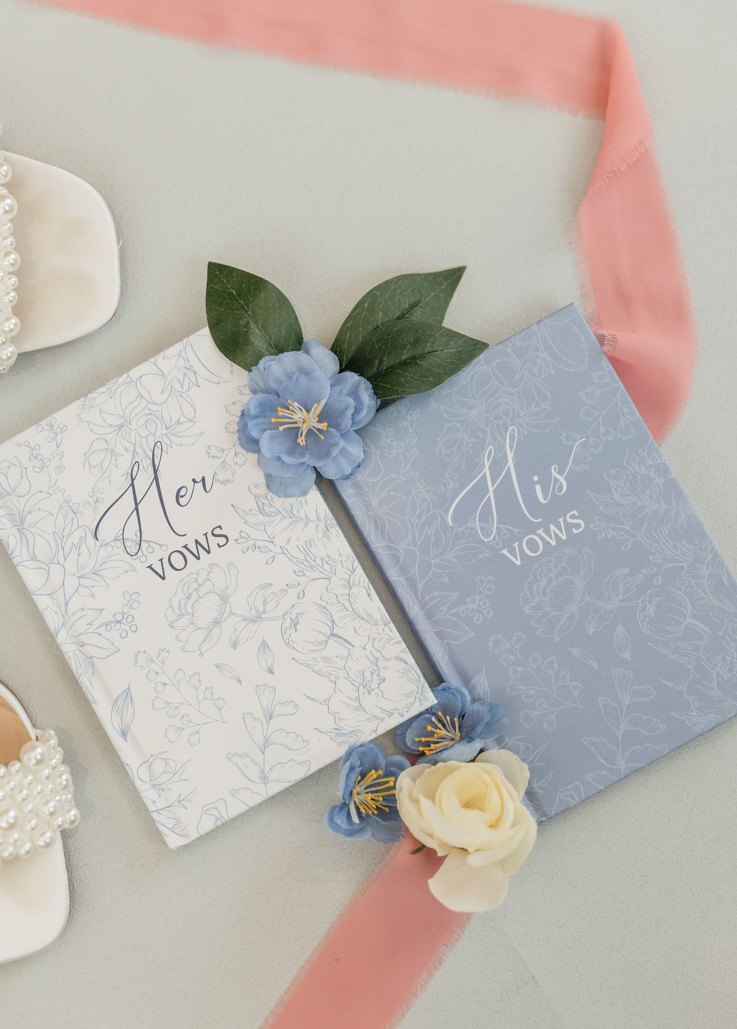 His and Her vows books in dusty blue and white with floral accents, pink ribbon, and pearl bridal shoes on gray background.