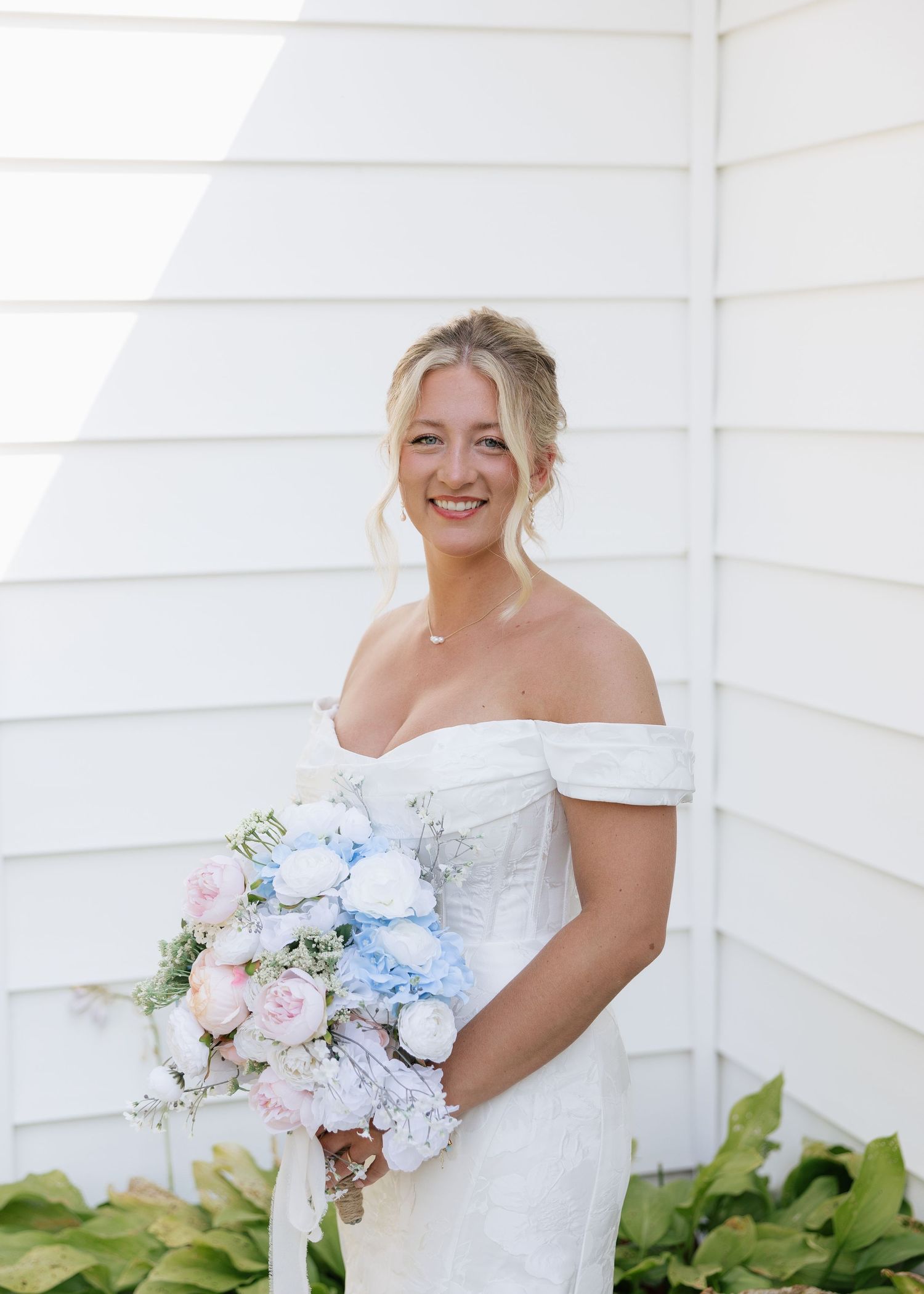 Classic bridal portrait with white off-shoulder dress and pastel bouquet against white siding.
