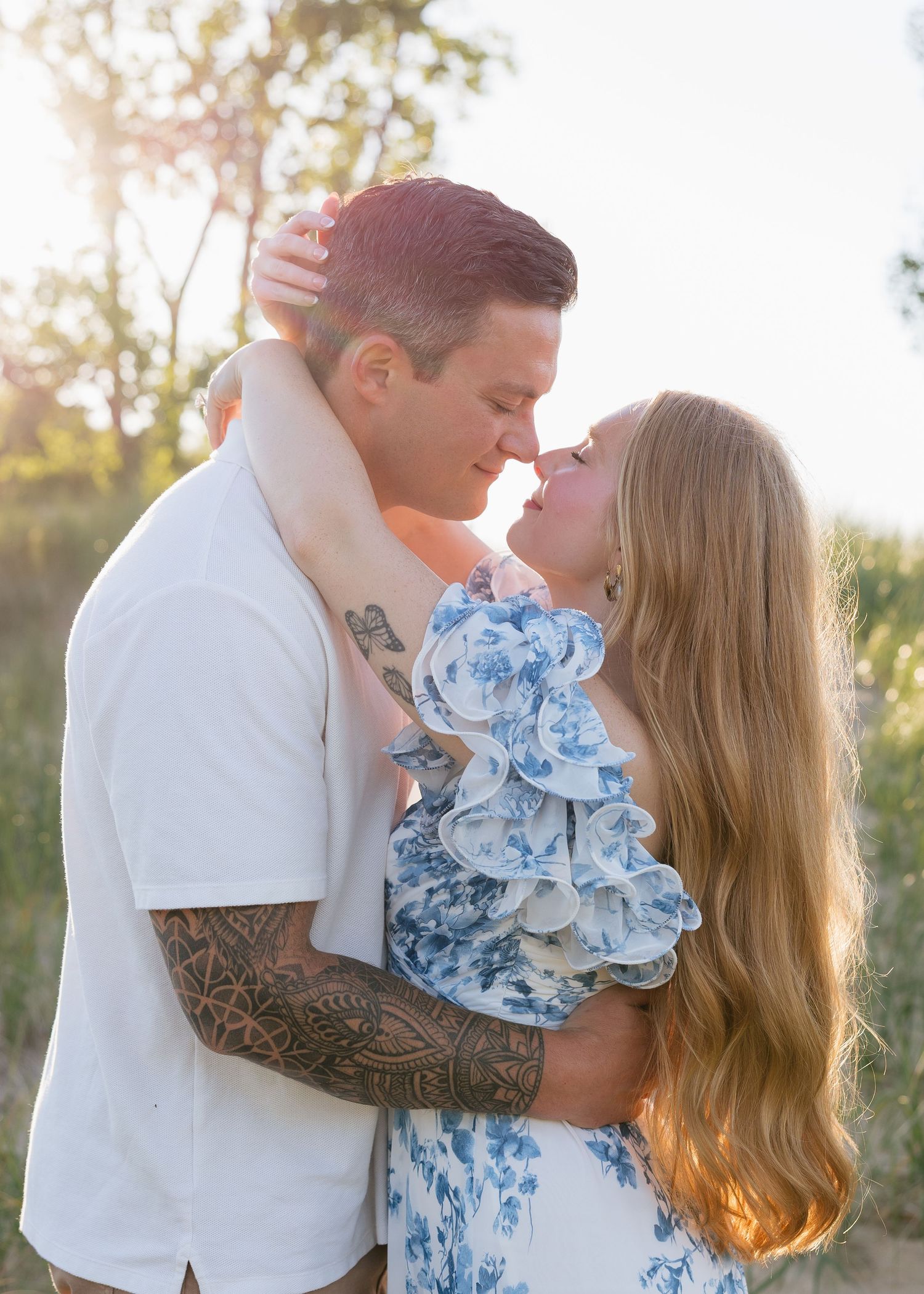 A couple shares a romantic almost-kiss outdoors at golden hour, man with tattoos in white shirt, woman in blue floral dress.