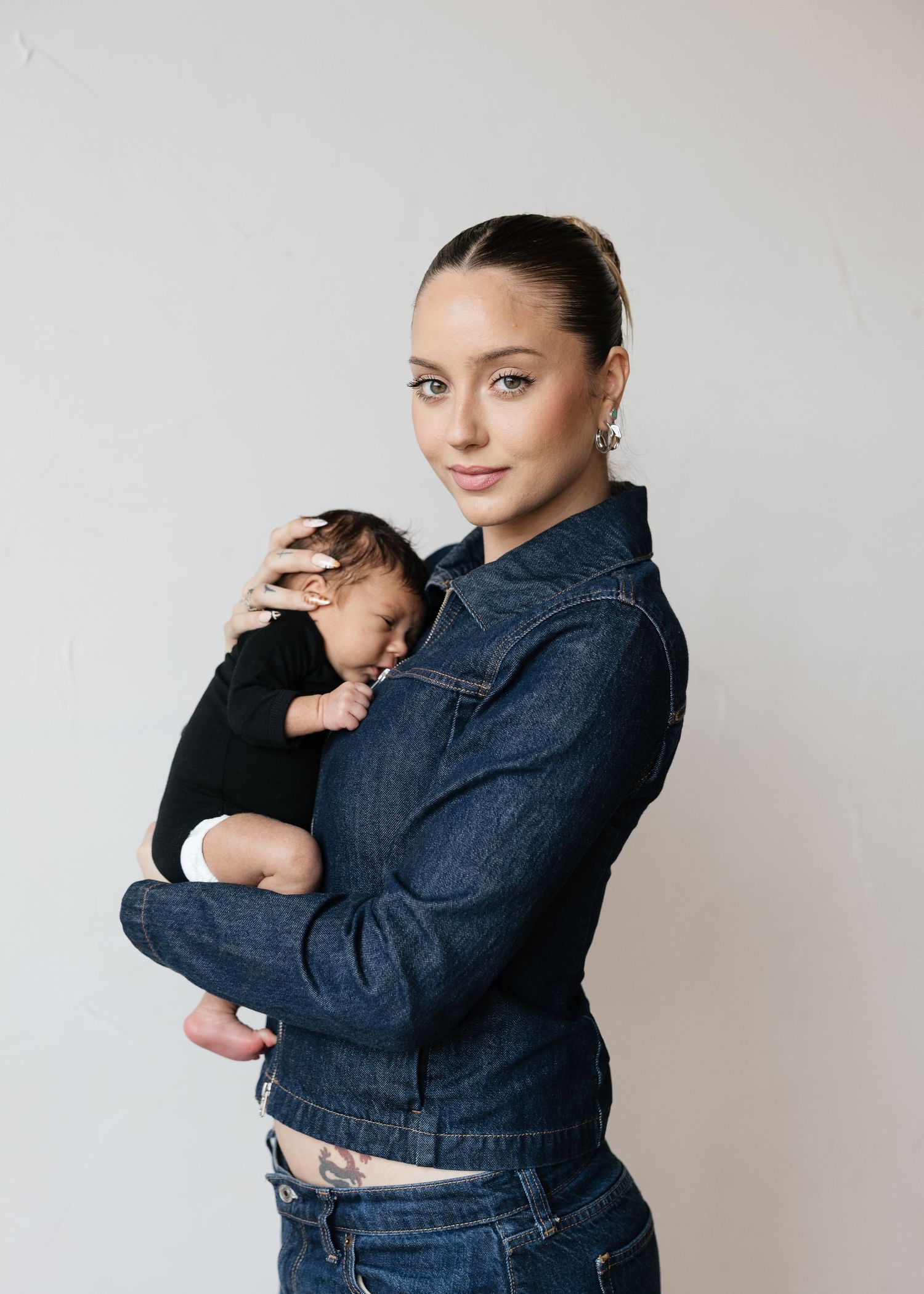 Young woman in denim jacket tenderly holds sleeping newborn baby against her chest, posing for a portrait.