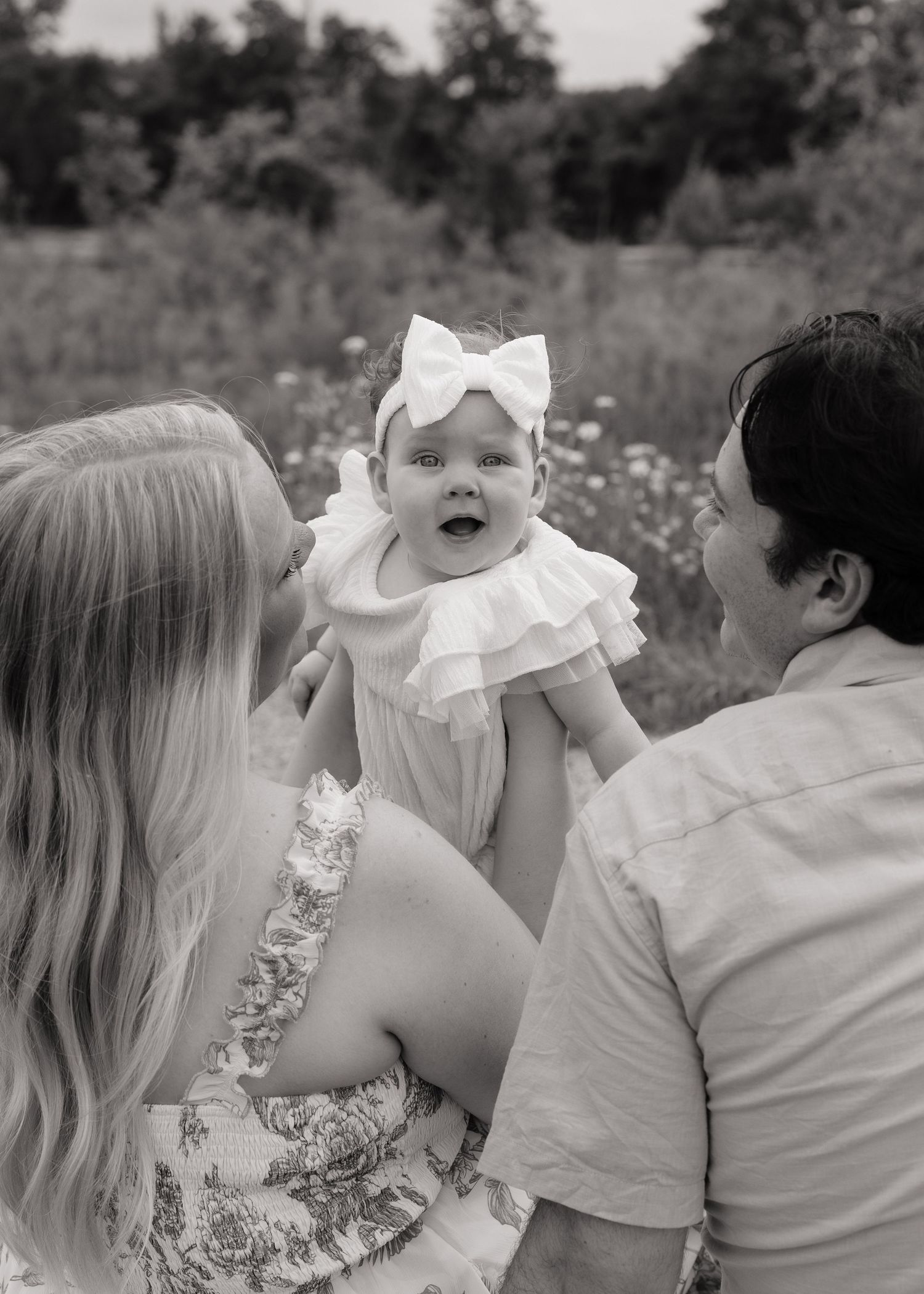 Black and white photo of parents lifting happy baby girl in white bow outdoors.
