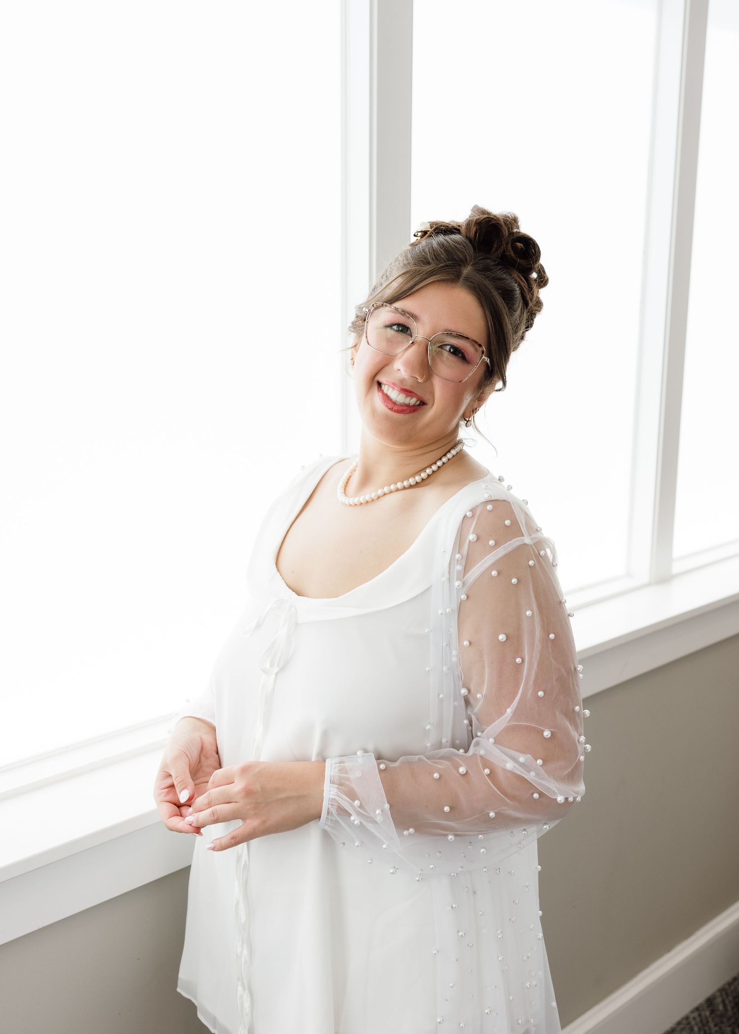 Smiling bride in white gown with sheer pearl sleeves and pearl necklace, posing by a bright window.