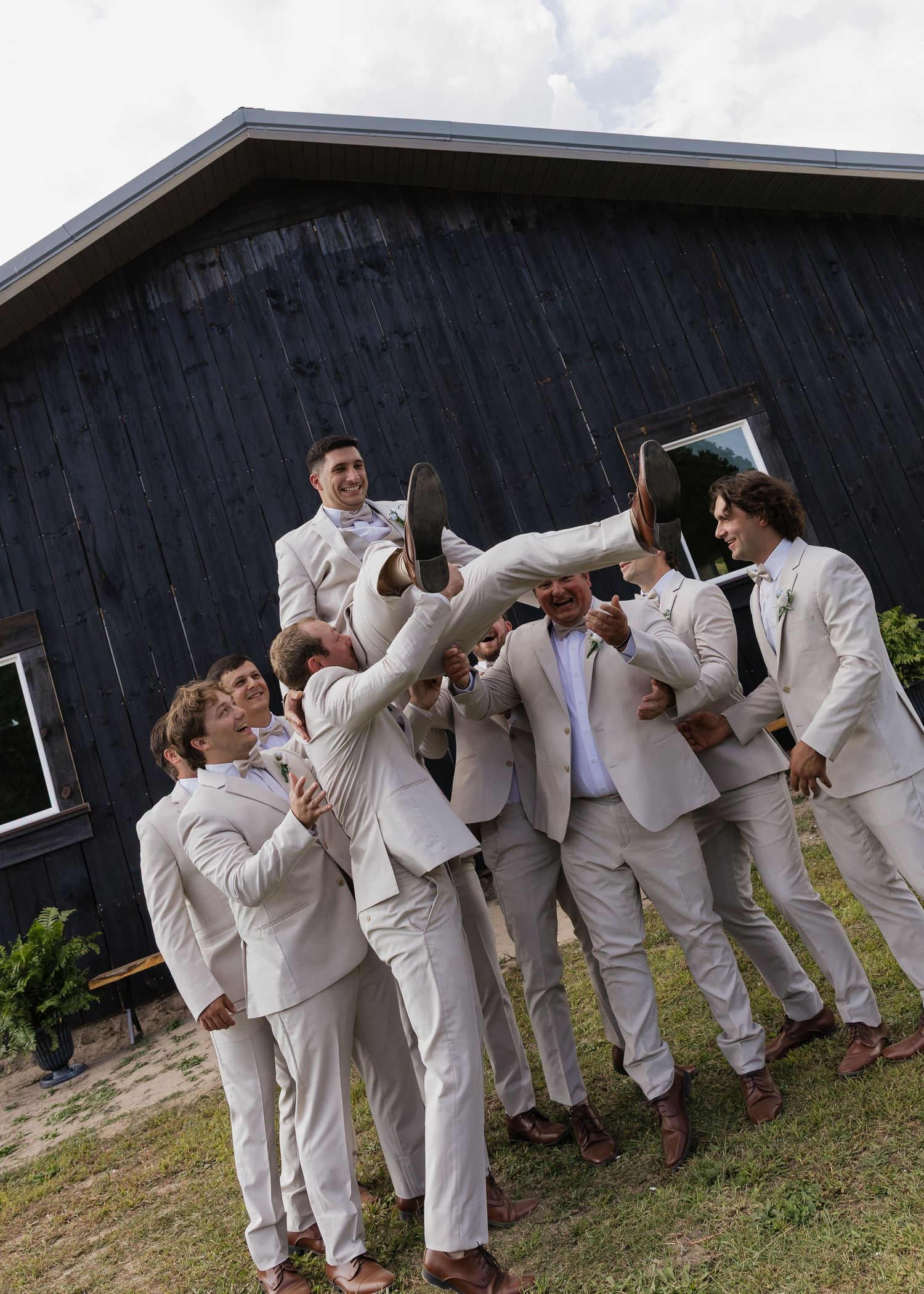 Groomsmen in white suits make playful poses against dark barn wall.