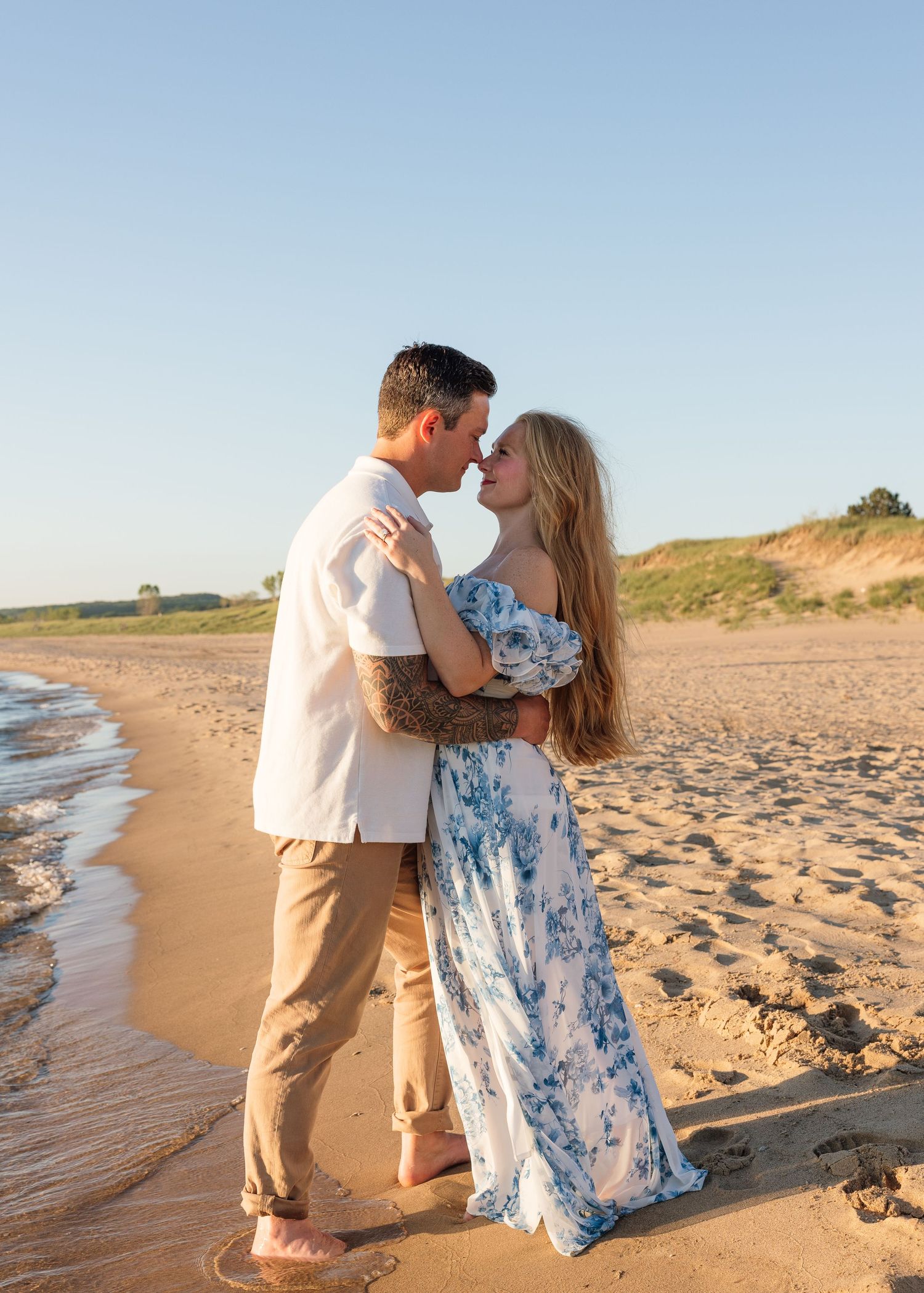 Couple sharing a romantic moment on a sandy beach at golden hour, woman in floral maxi dress, man with tattoos.