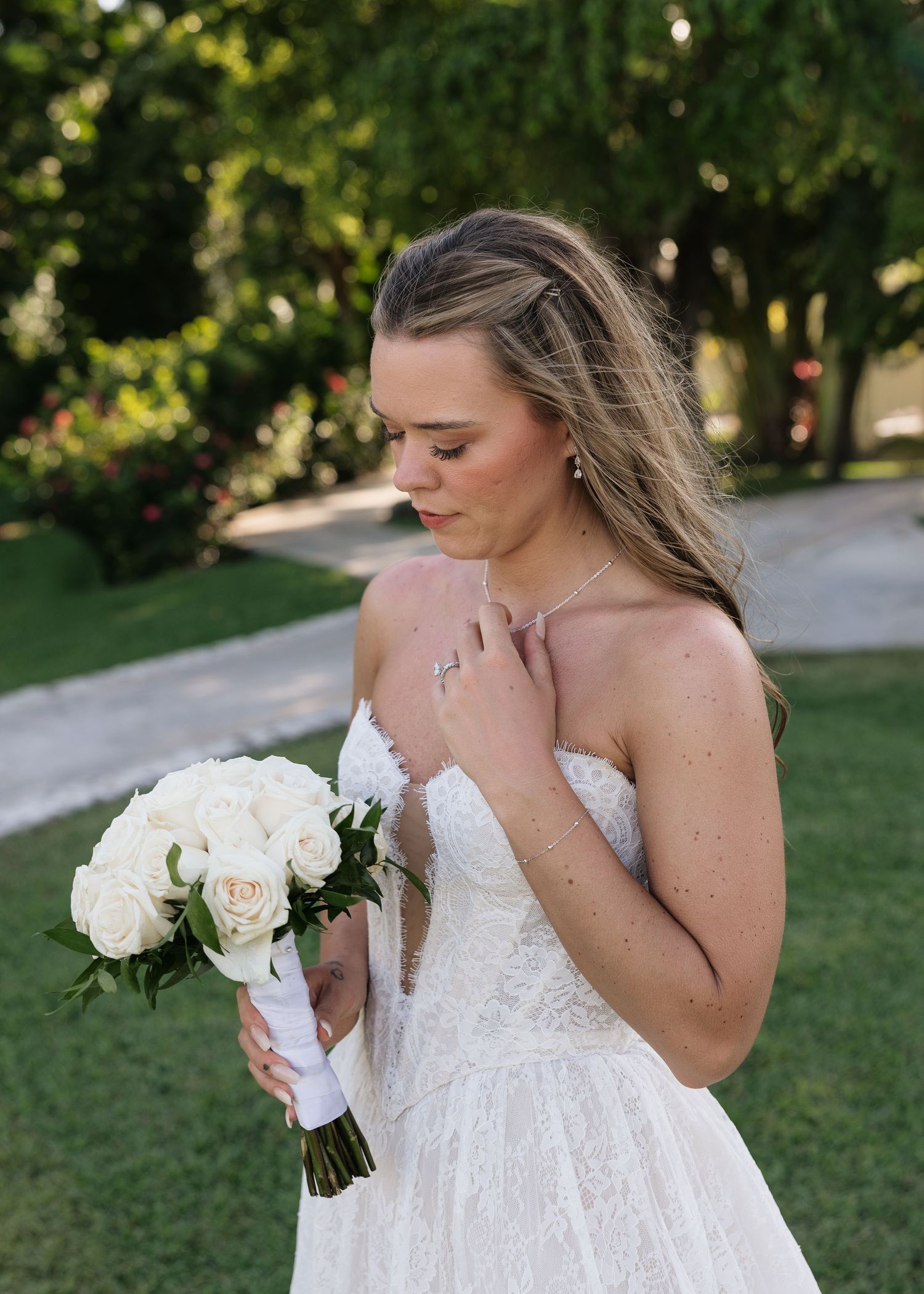 Bride in lace gown holds white rose bouquet outdoors, looking down gracefully on her wedding day.