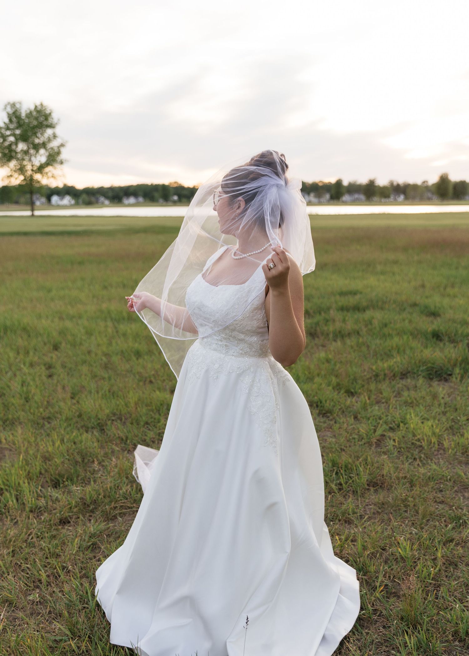 Bride in white wedding gown and veil standing in green field at sunset, holding veil over face.