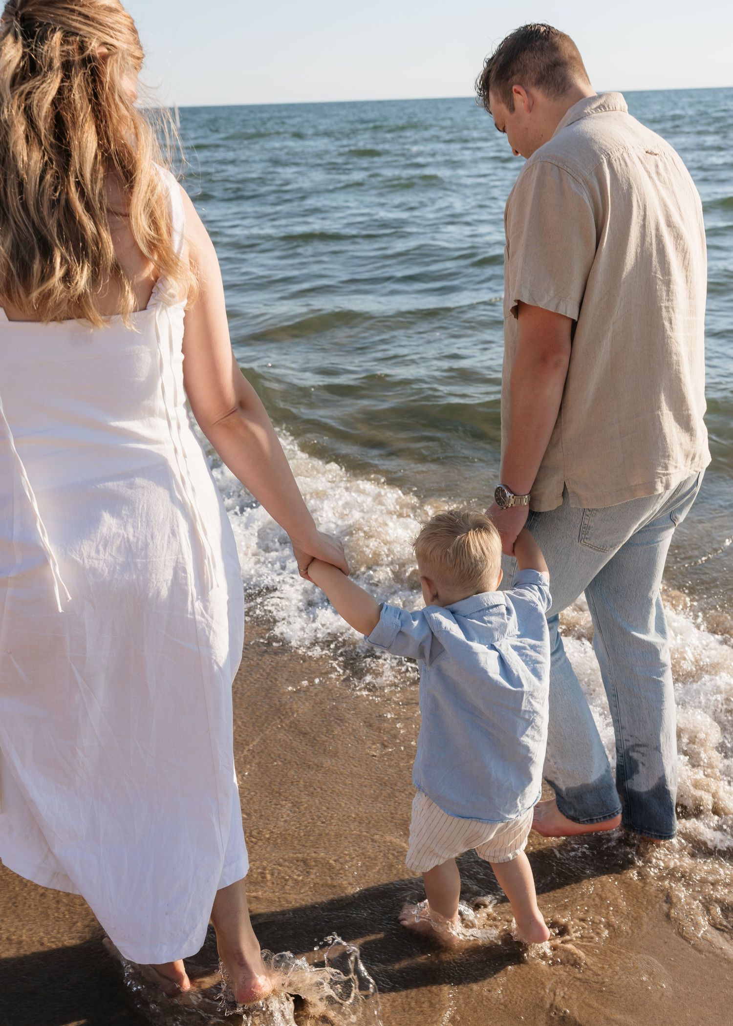 Family of three holding hands at the ocean shore, parents and toddler standing at water's edge on a sunny day.