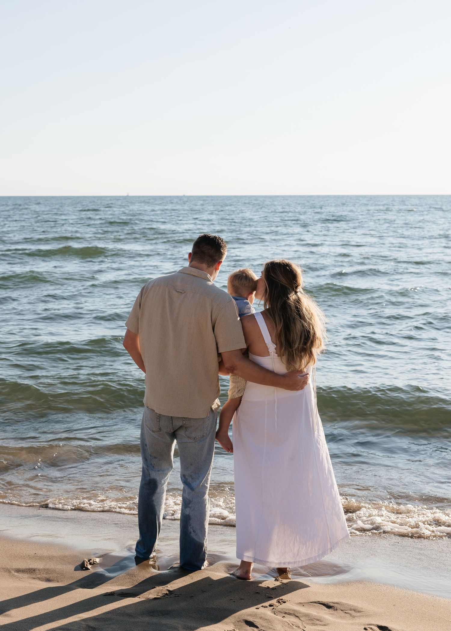 Family of three standing on sandy beach facing ocean waves, mother holding baby in white dress beside father in casual attire.