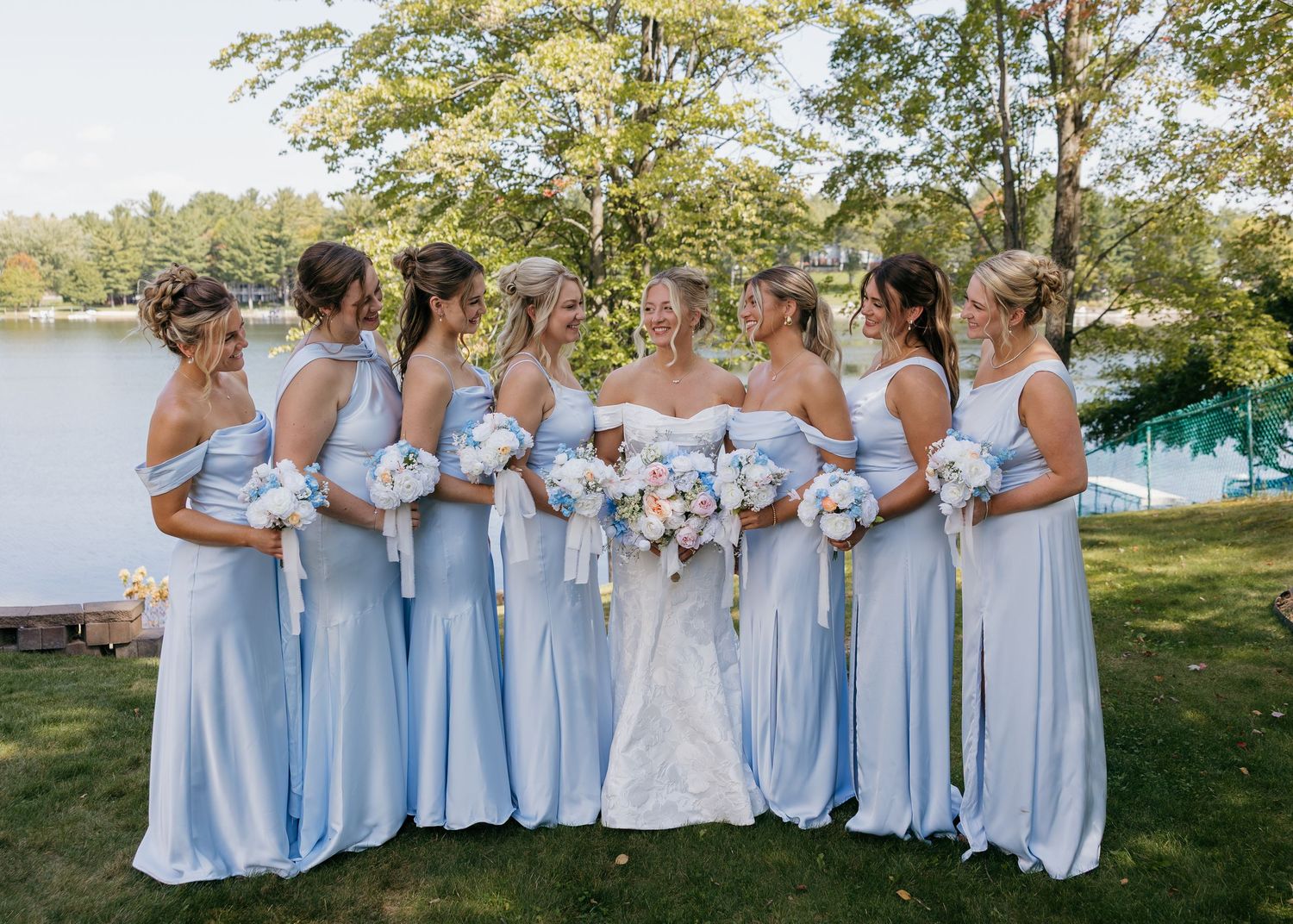 Wedding party in light blue bridesmaid dresses posing by lake with trees in background.