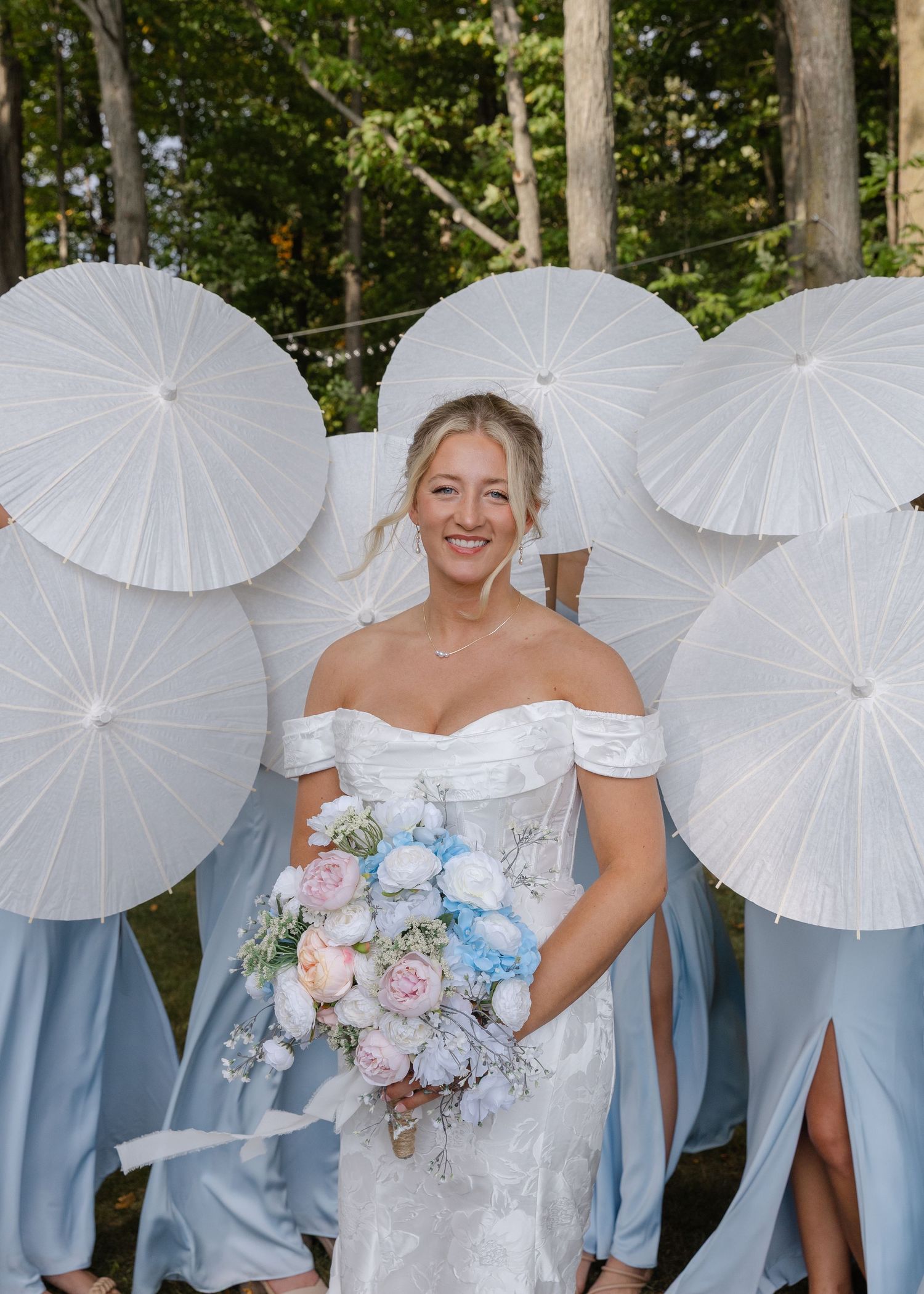 Wedding party poses with white parasols and light blue dresses against a wooded backdrop.