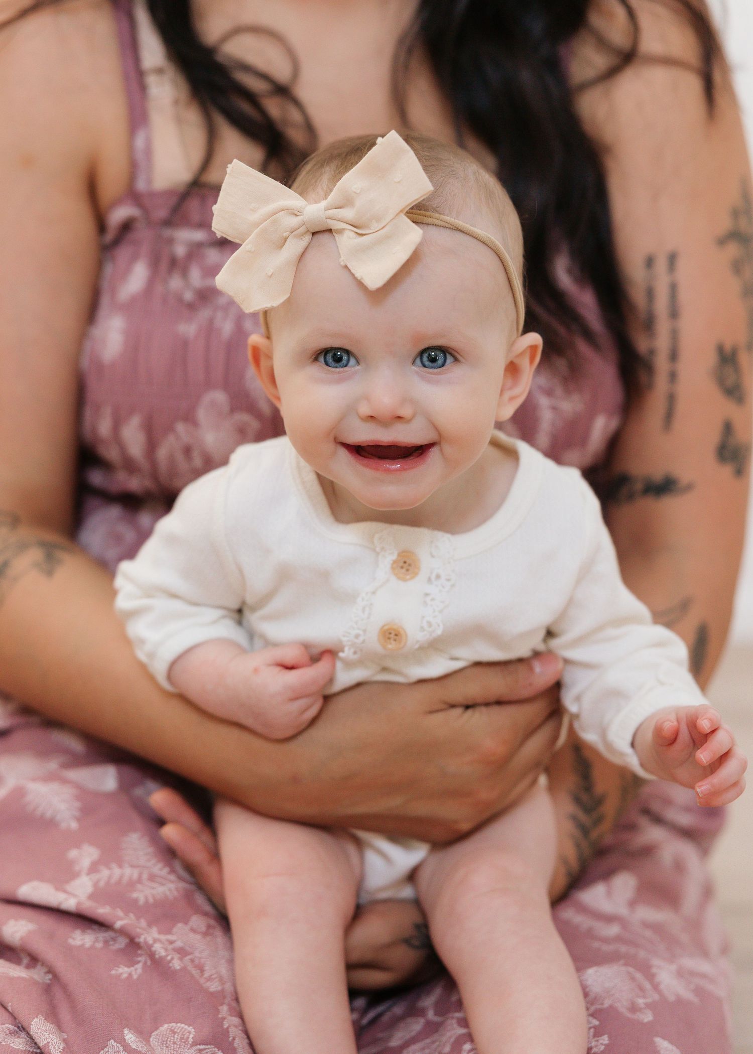 Smiling baby girl with blue eyes wearing a cream outfit and bow headband, held by a tattooed woman in a floral dress.