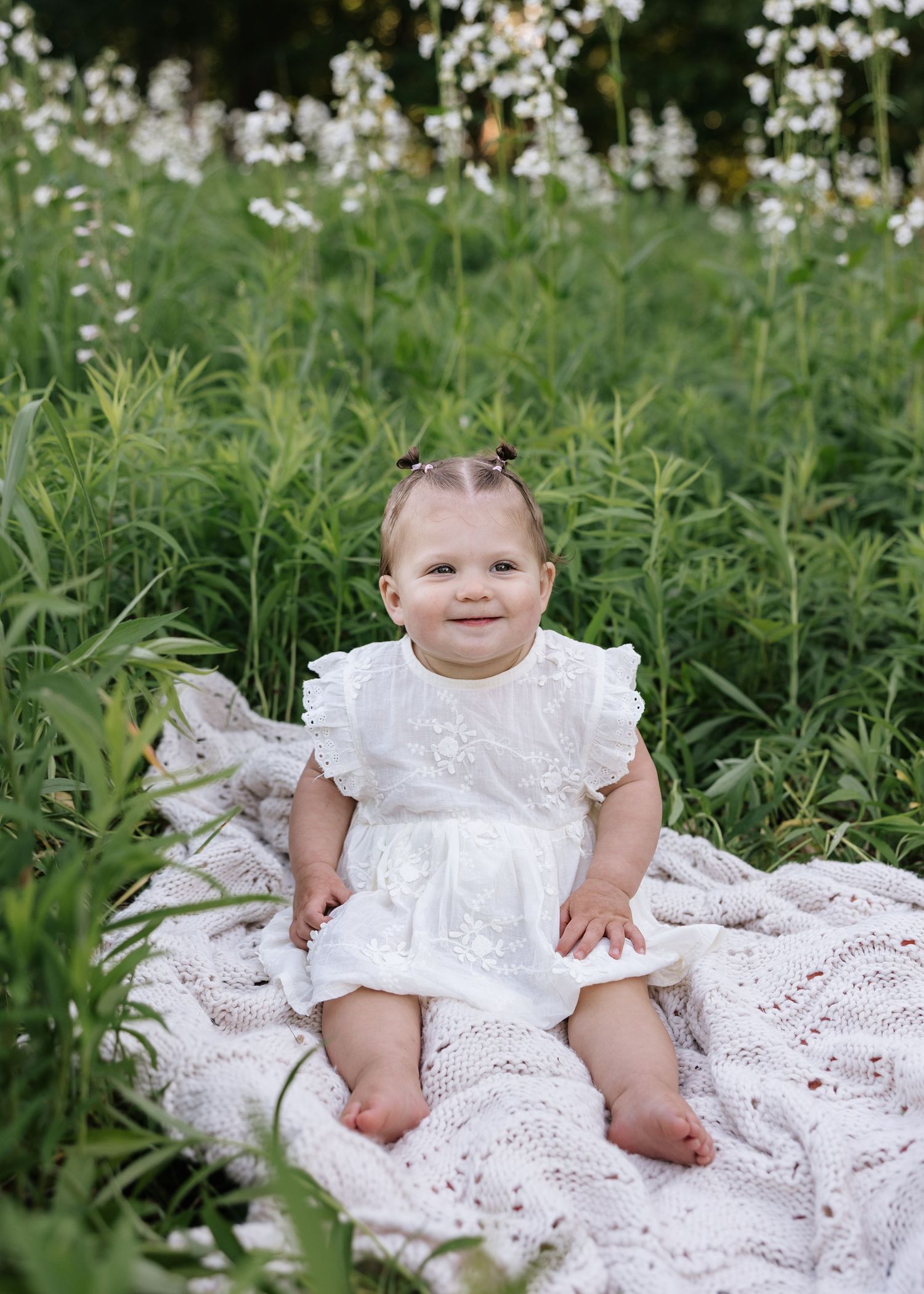 A smiling baby girl in a white dress sits on a lace blanket surrounded by lush green wildflowers.