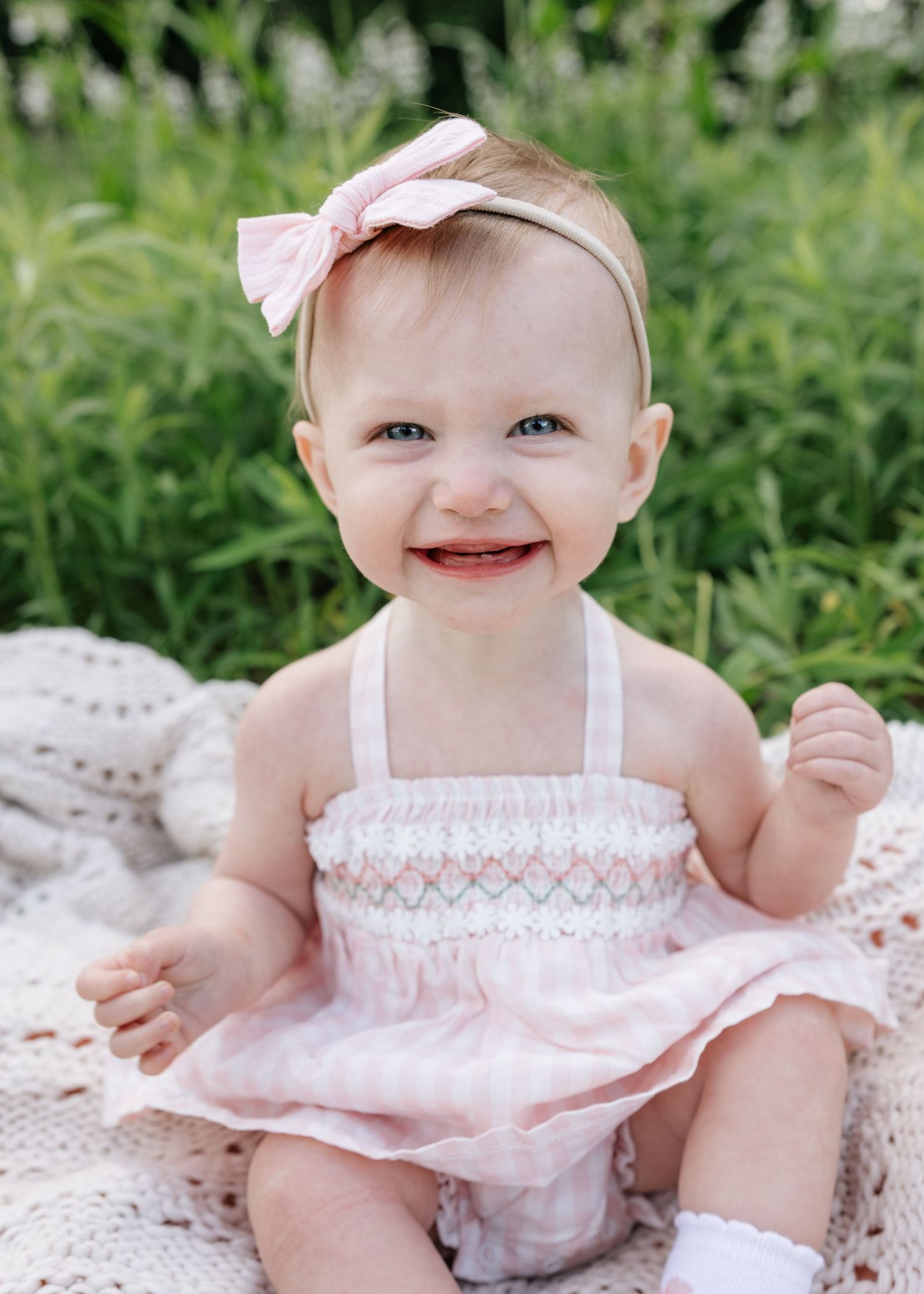 Smiling baby girl in pink smocked romper and bow headband sitting on blanket outdoors with green foliage background.