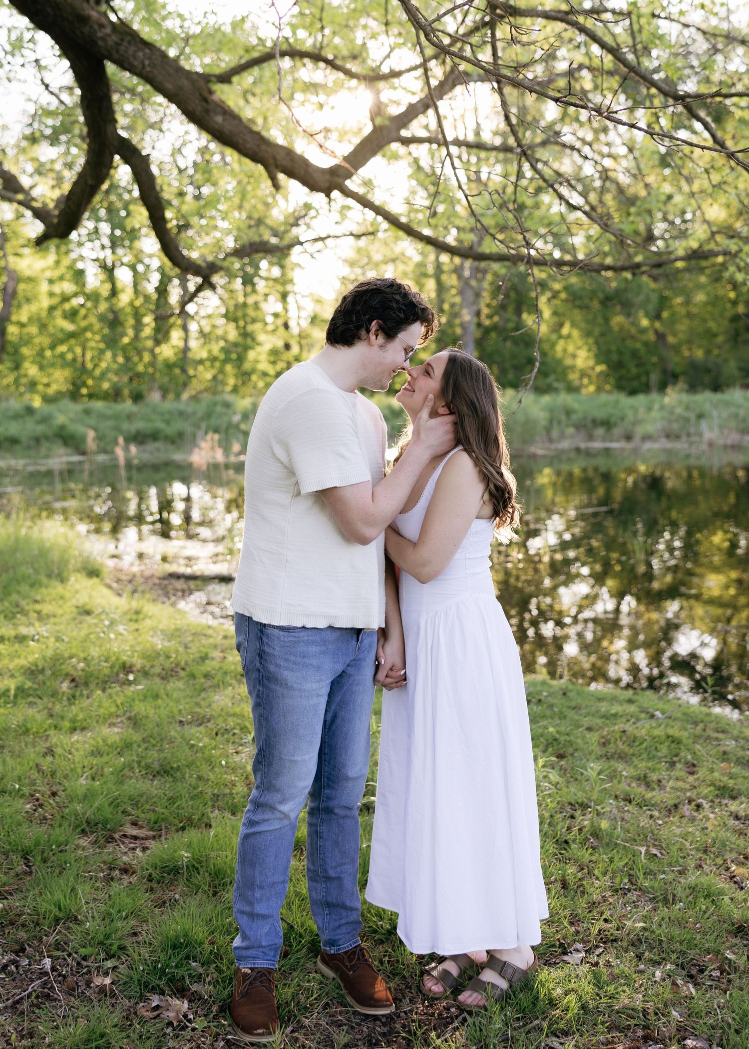 Couple sharing a romantic moment by a serene pond, surrounded by lush greenery in golden hour light.