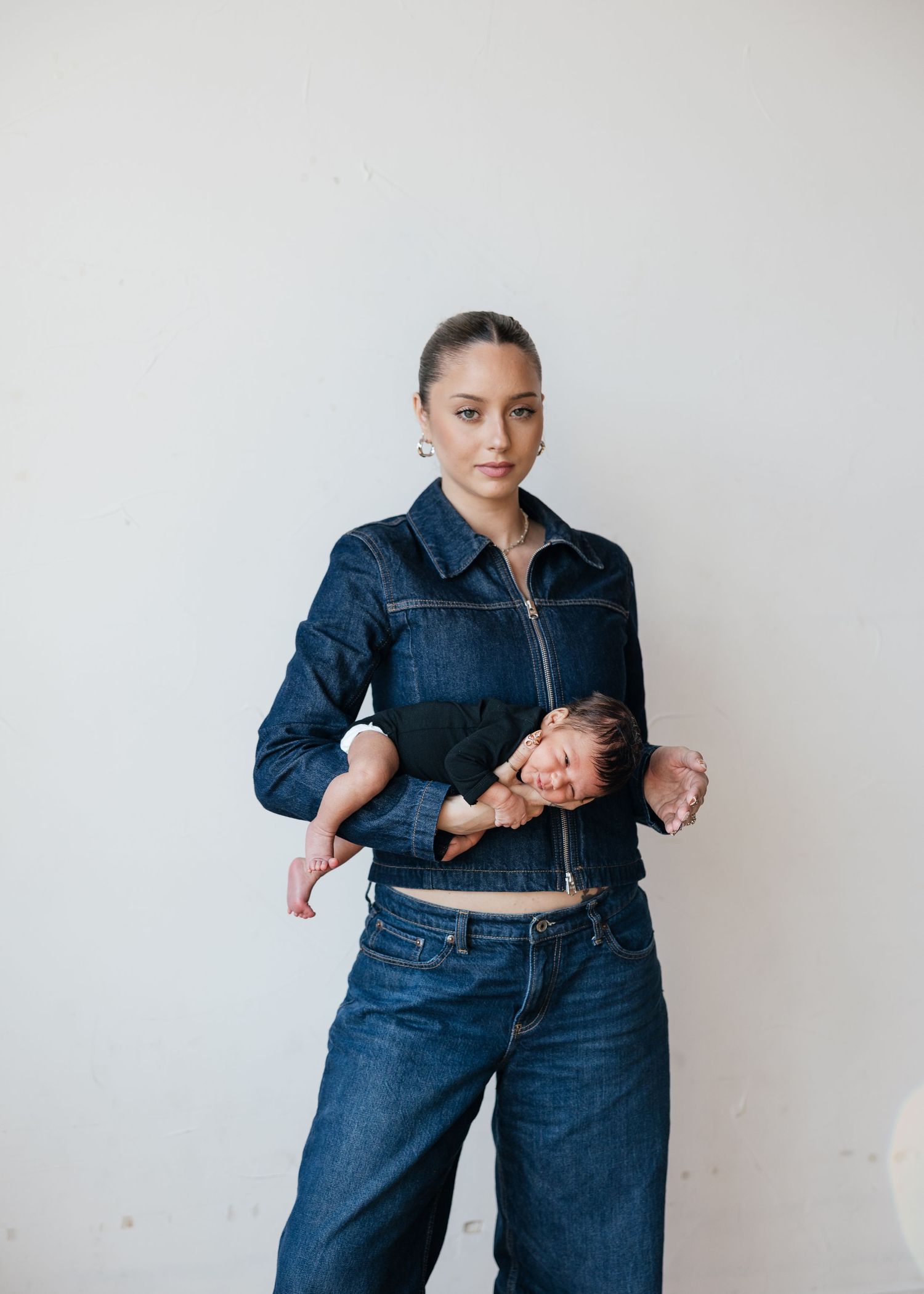 Woman in dark denim jacket and jeans holds a baby against her chest while standing against a white wall.