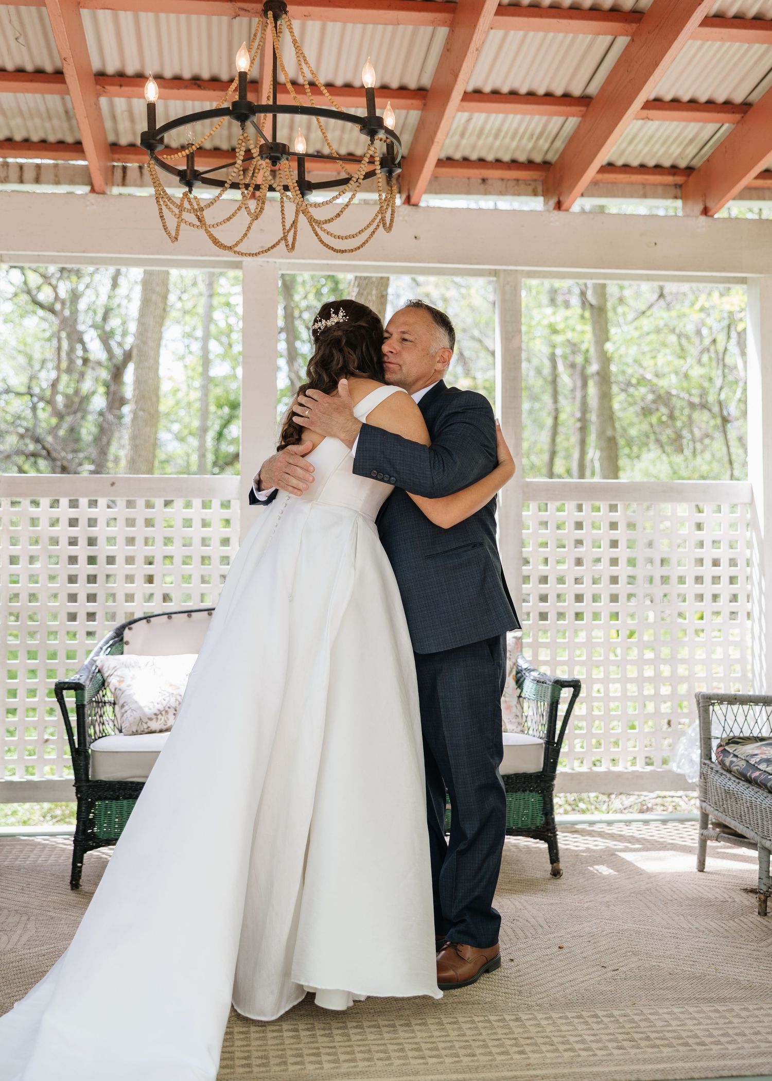 Bride in white gown shares emotional first look hug with father on decorated porch at outdoor wedding venue.