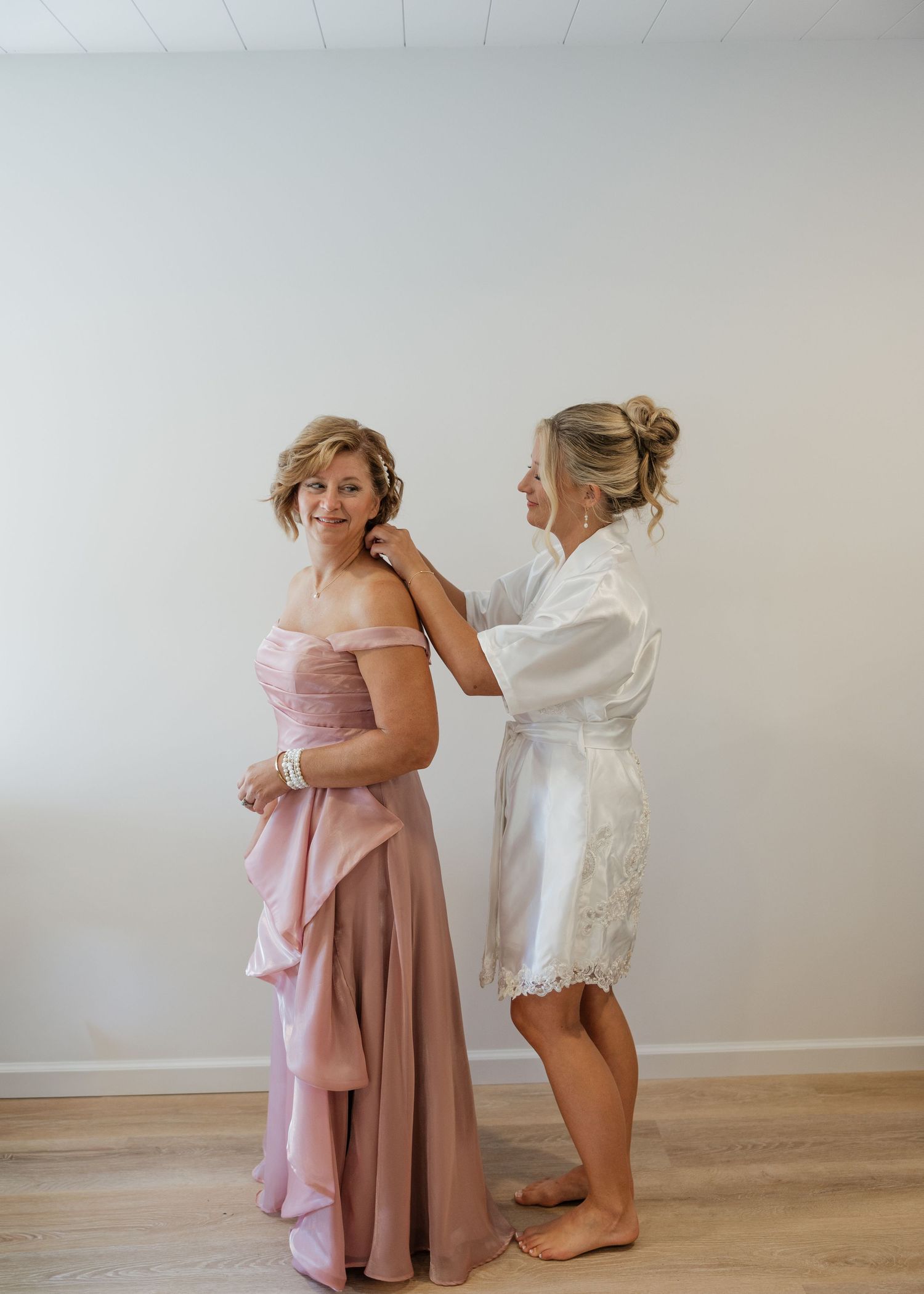 Two people getting ready for a wedding, with one wearing a pink dress and the other in a white robe.