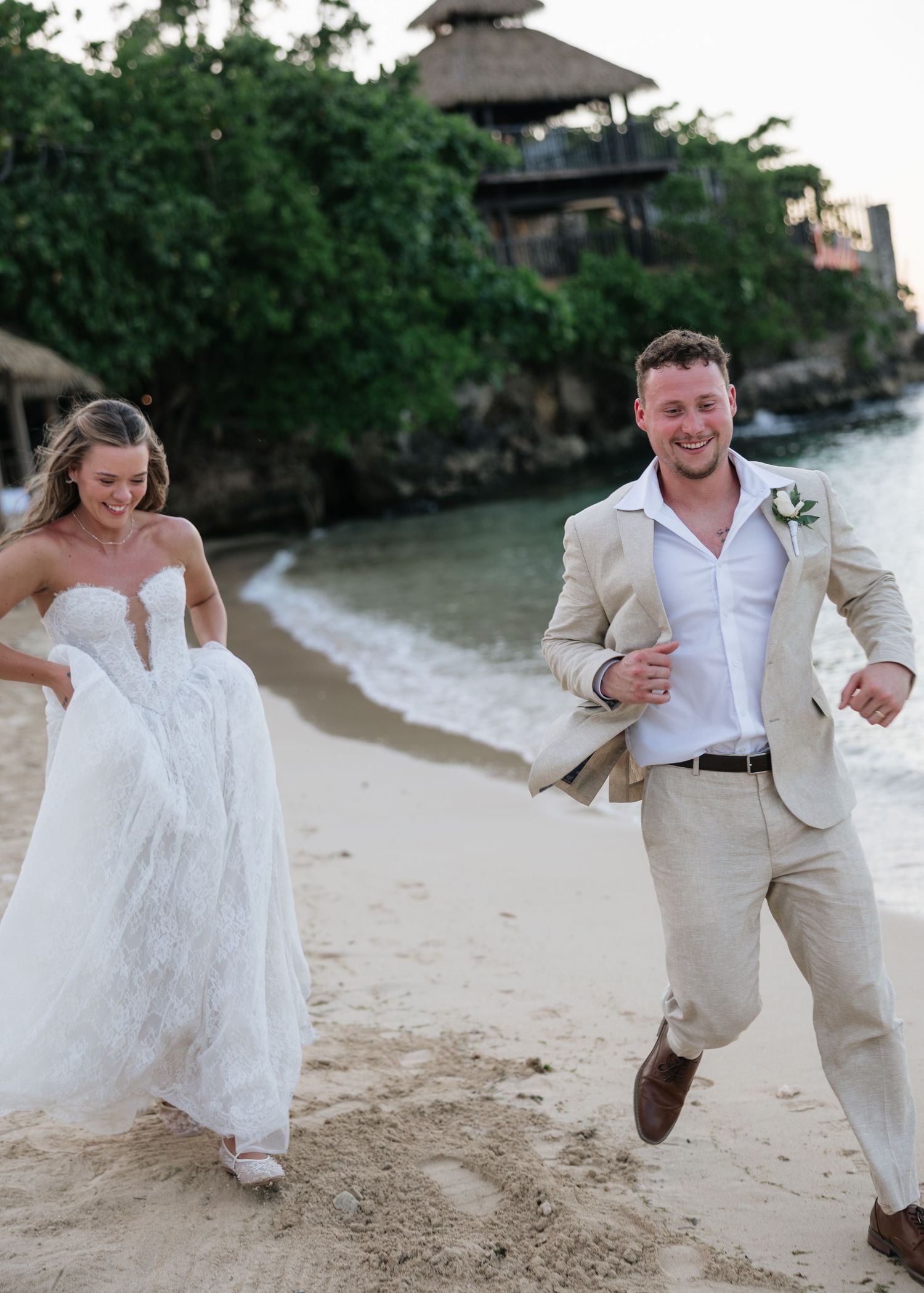 Bride and groom running joyfully on a sandy beach in wedding attire, with tropical resort scenery in the background.