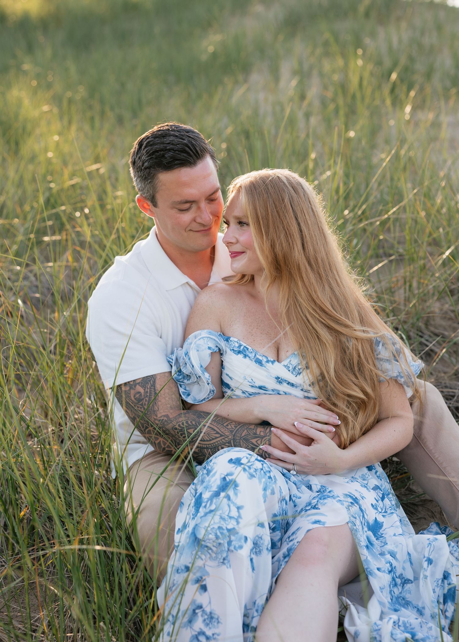 Couple sitting in tall grass at golden hour, woman in blue floral dress, man in white shirt with tattoos, embracing tenderly.