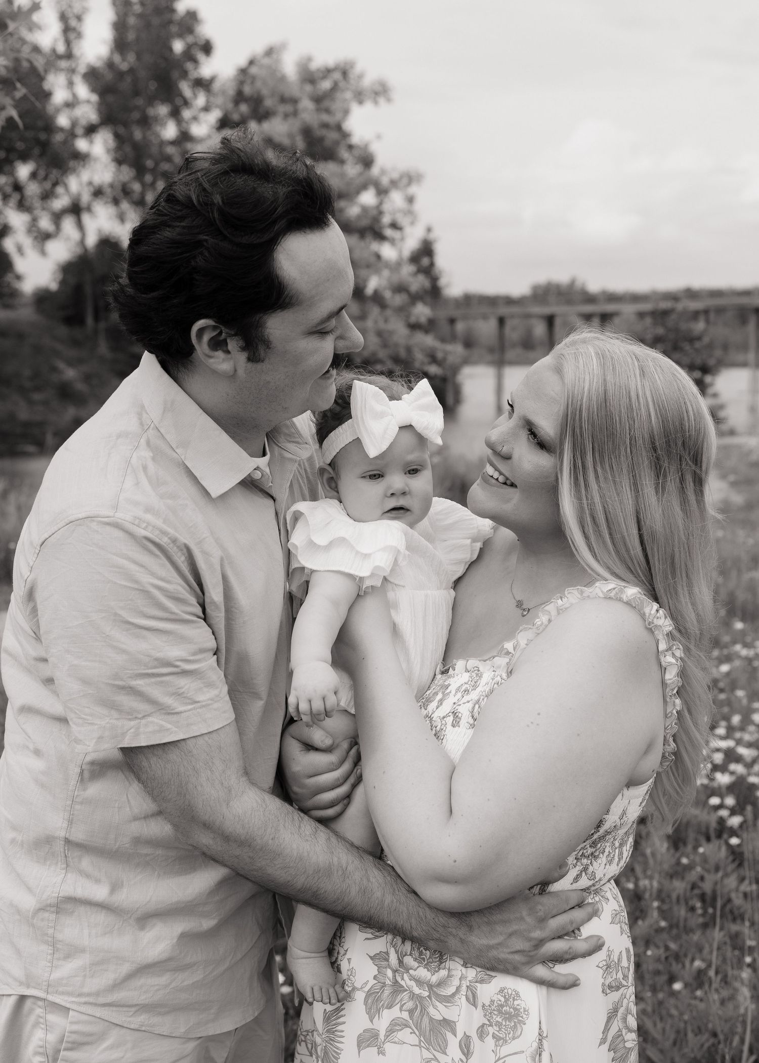 Black and white photo of a couple smiling at each other while holding their baby girl with a bow in an outdoor setting.